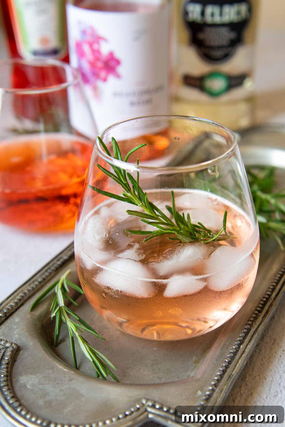 A vibrant rosé spritzer cocktail in a glass, with bottles for making the drink subtly blurred in the background, set on a bright, inviting surface.
