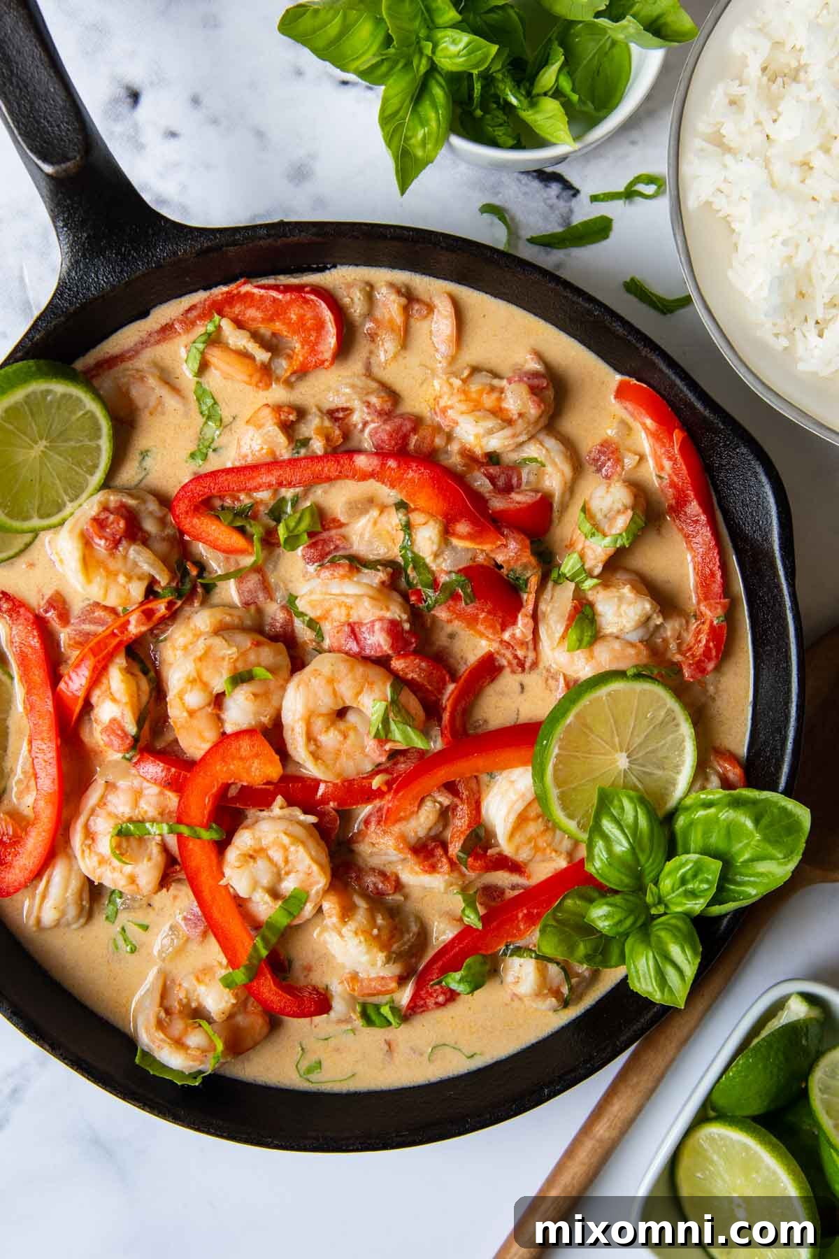 Overhead shot of completed creamy coconut shrimp in a skillet, garnished with fresh herbs, with a bowl of limes and a side of jasmine rice.