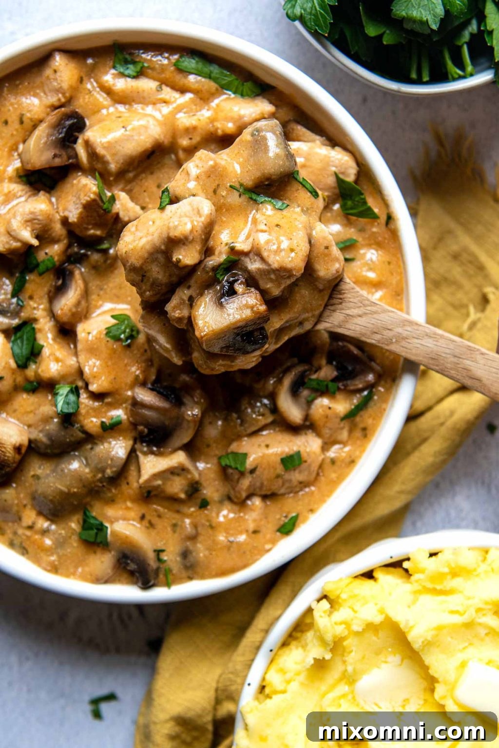 A wooden spoon carefully lifts a serving of creamy chicken stroganoff from a pristine white bowl, showcasing the rich sauce and tender chicken.