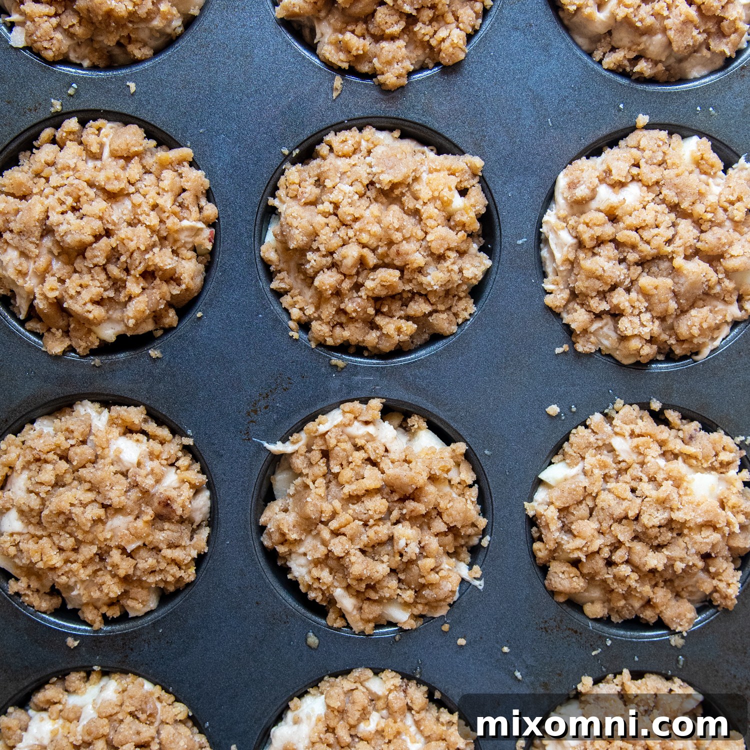 Unbaked gluten-free apple muffins in a tin, topped with cinnamon brown sugar crumb topping, ready for the oven.