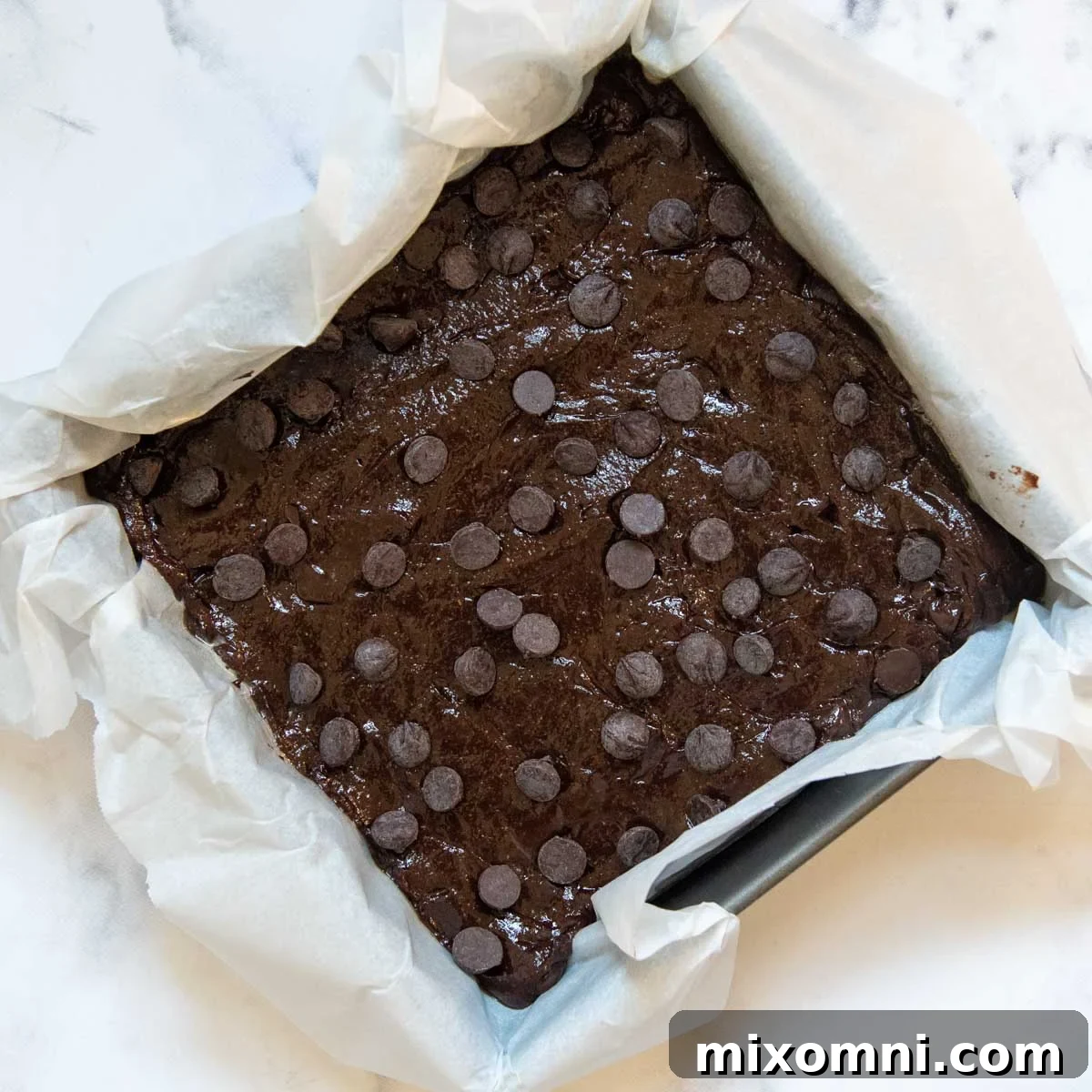 Raw brownie batter spread into a parchment-lined baking pan, topped with an extra sprinkle of chocolate chips, ready for the oven.