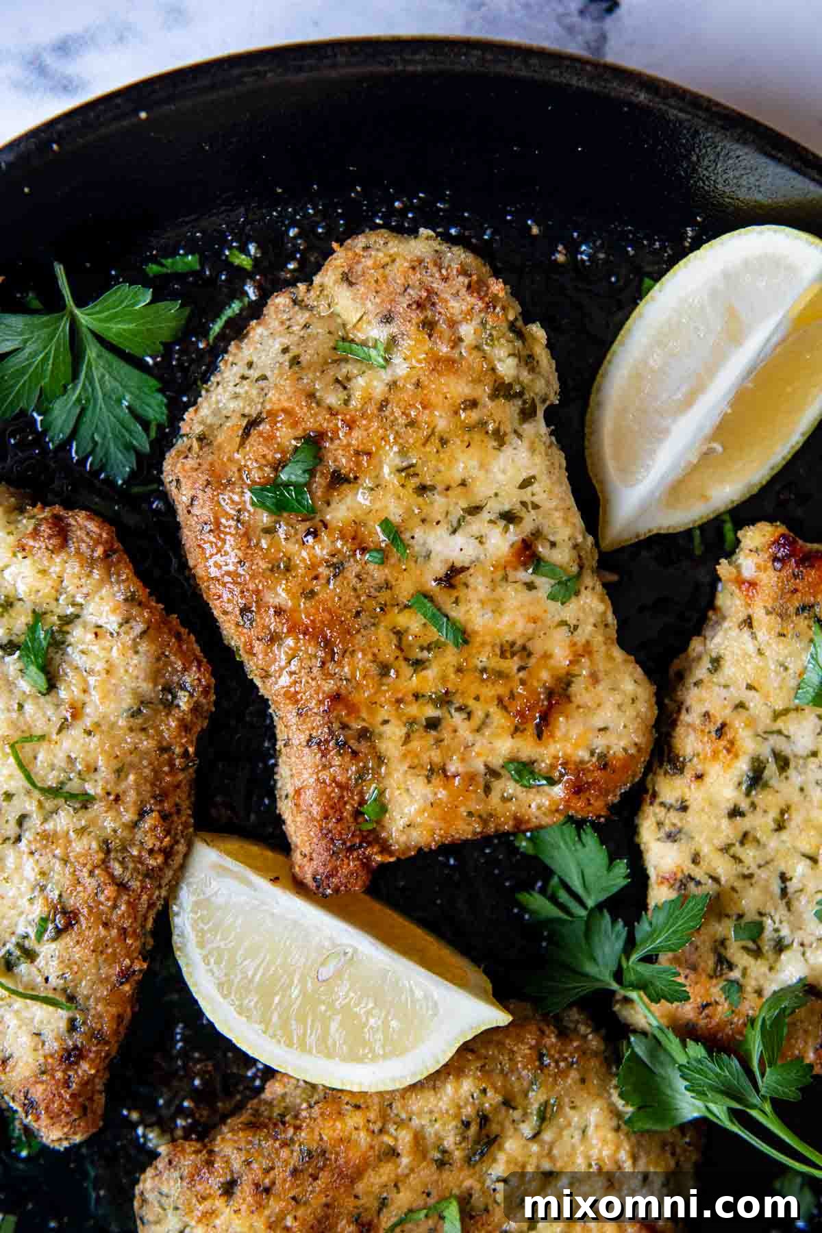Overhead shot of freshly baked breaded pork chops in a black pan, garnished with lemon wedges and fresh herbs.