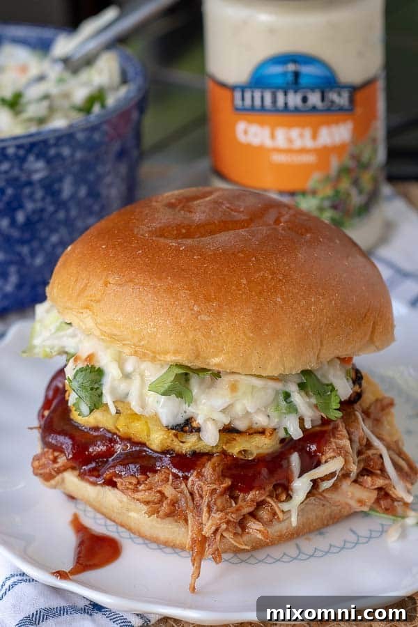a BBQ pulled chicken sandwich on a plate with coleslaw, BBQ sauce, and grilled pineapple