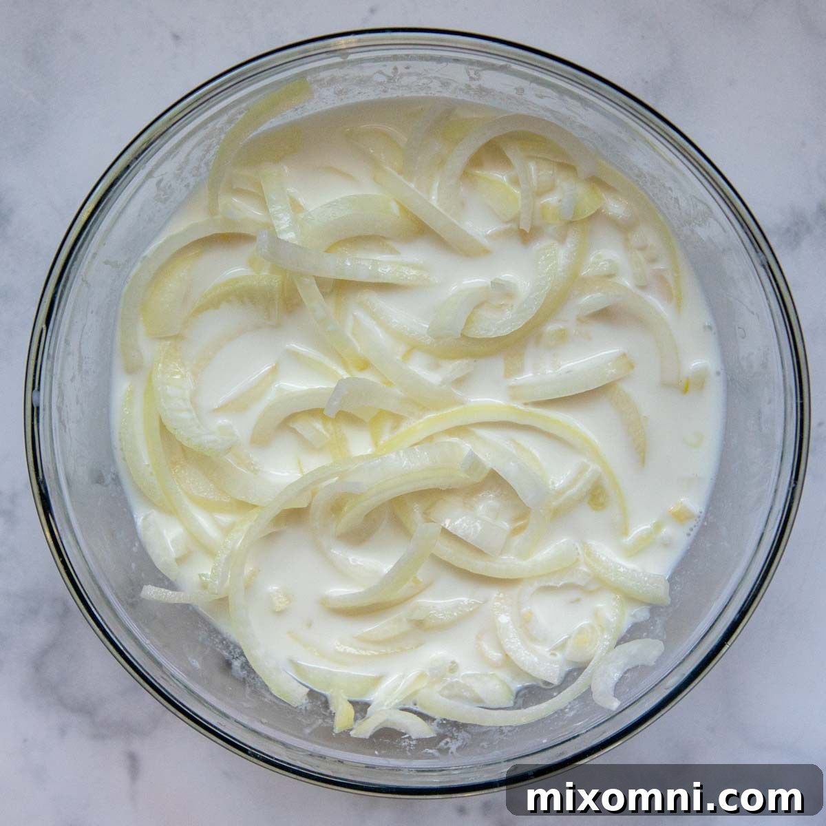 Thinly sliced raw onions gently soaking in a bowl of buttermilk, preparing them for the frying process.