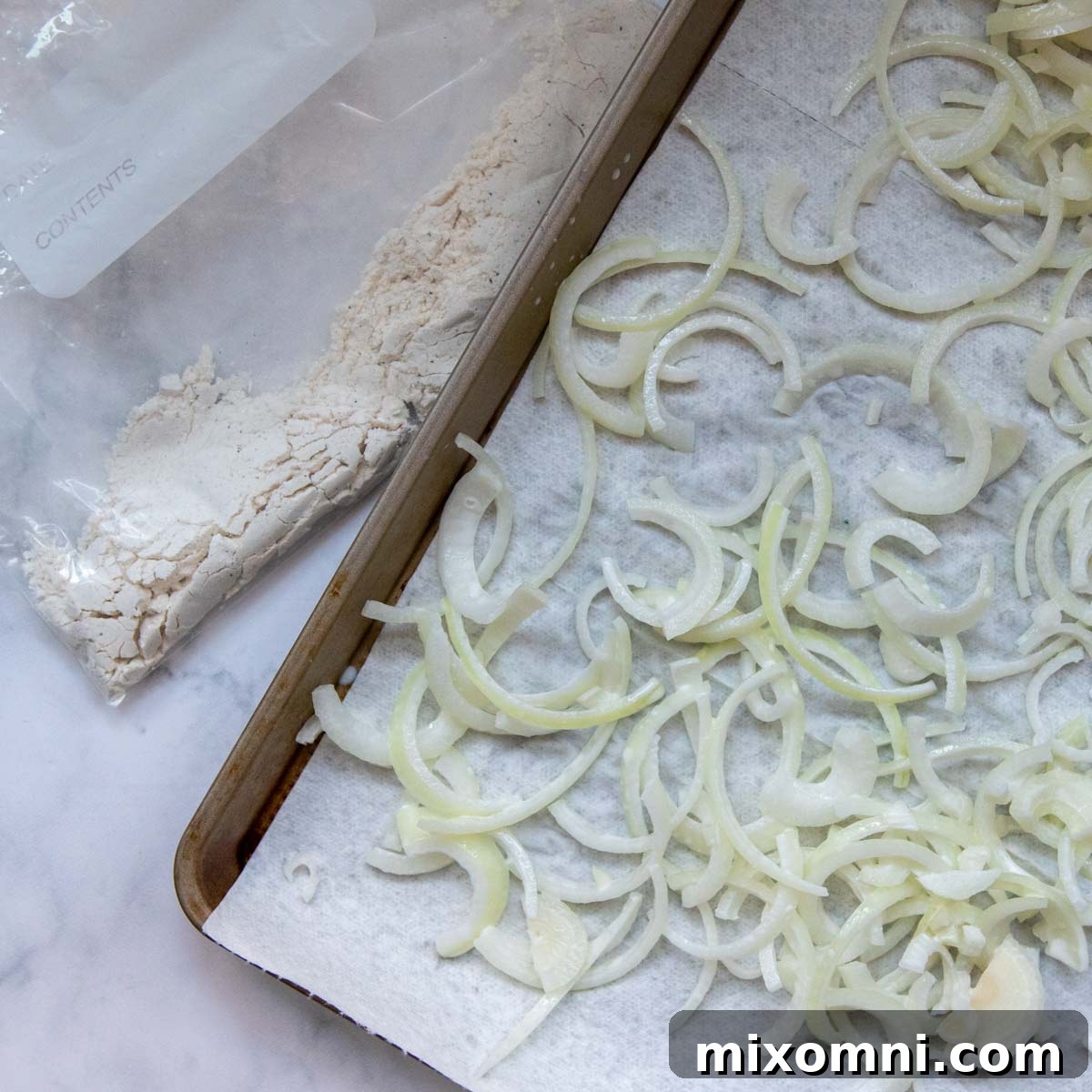 Onions, lightly damp from their buttermilk soak, resting on a paper-towel-lined baking sheet to absorb excess moisture before coating.