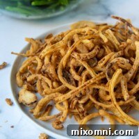 A plate of golden, crispy homemade French fried onions with a bowl of green bean casserole in the background.