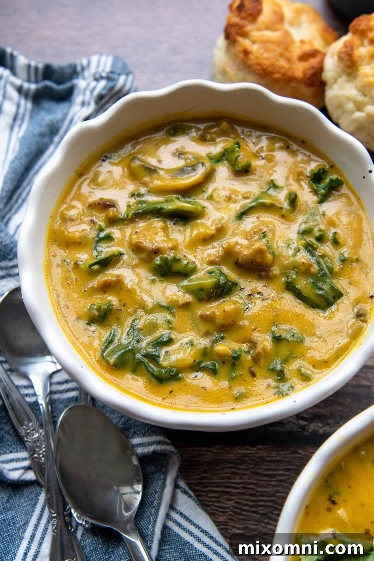 A steaming bowl of creamy pumpkin sausage soup, garnished with fresh herbs, served on a rustic wooden table with a blue napkin and crusty biscuits in the background. Perfect for a cozy fall meal.