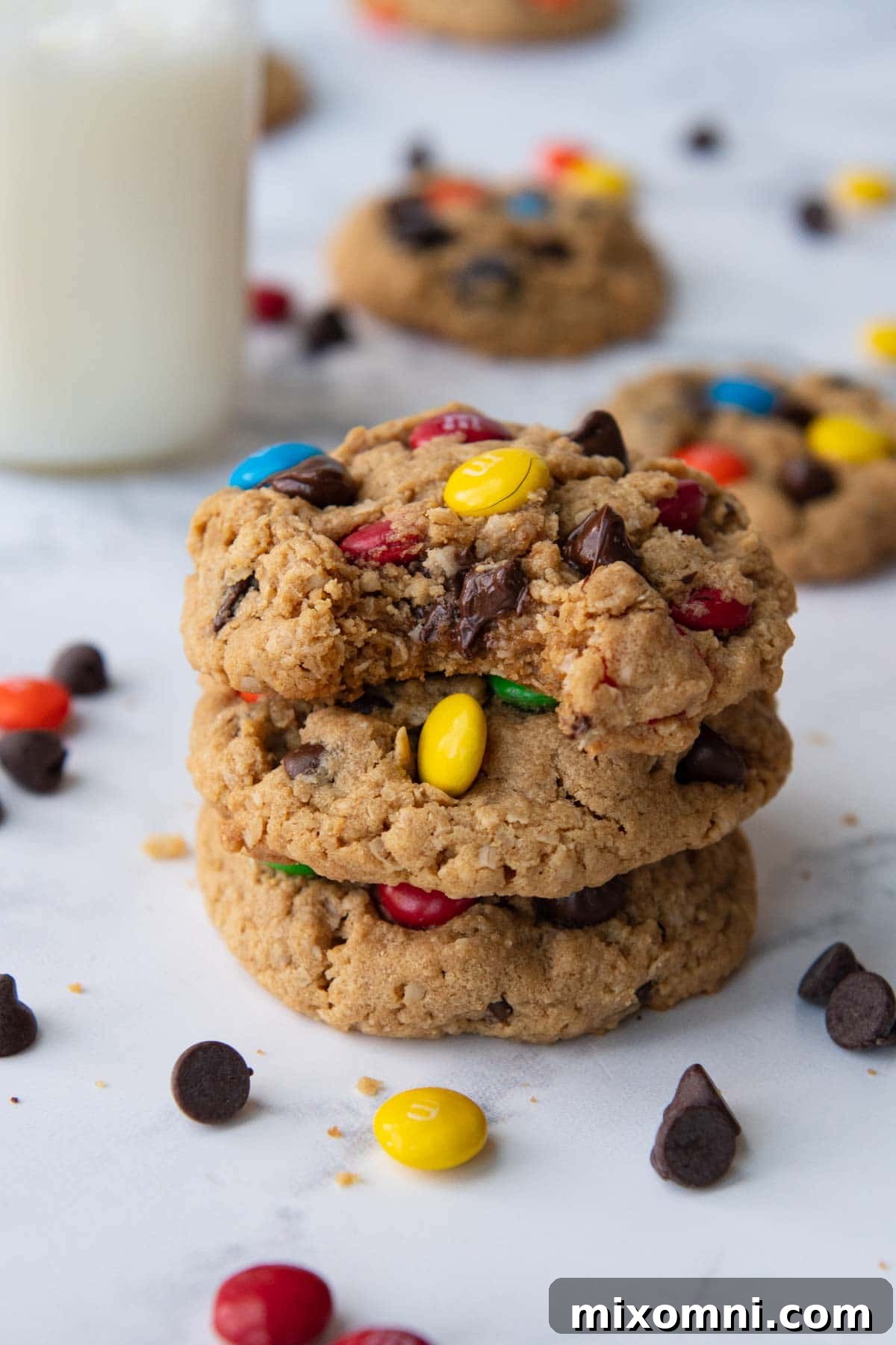 A stack of three golden-brown gluten-free monster cookies, with a bite taken out of the top one, revealing gooey chocolate chips and M&M candies inside.