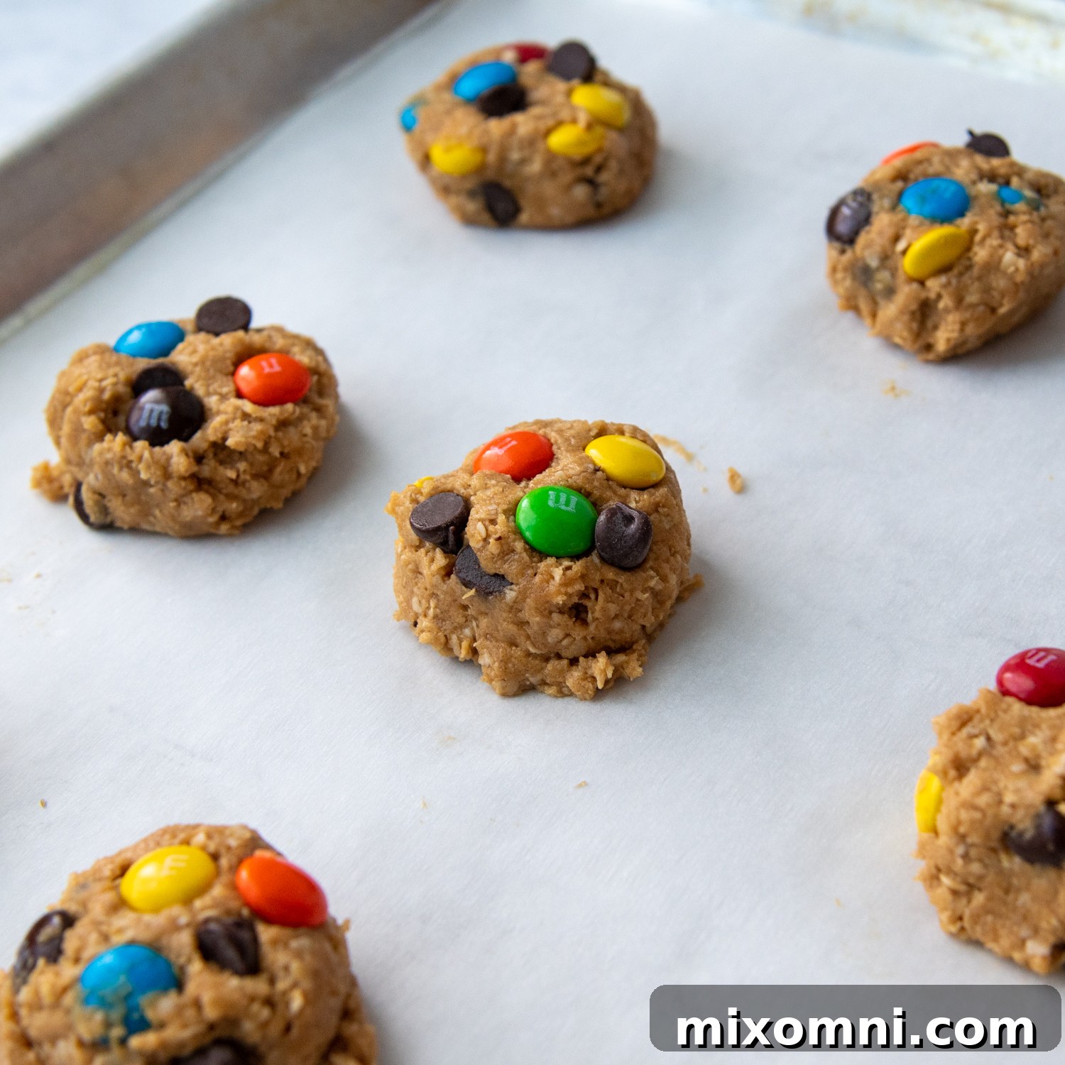 Unbaked, perfectly shaped monster cookie dough balls with extra chocolate chips and M&M's pressed on top, arranged on a parchment-lined baking sheet, ready for the oven.