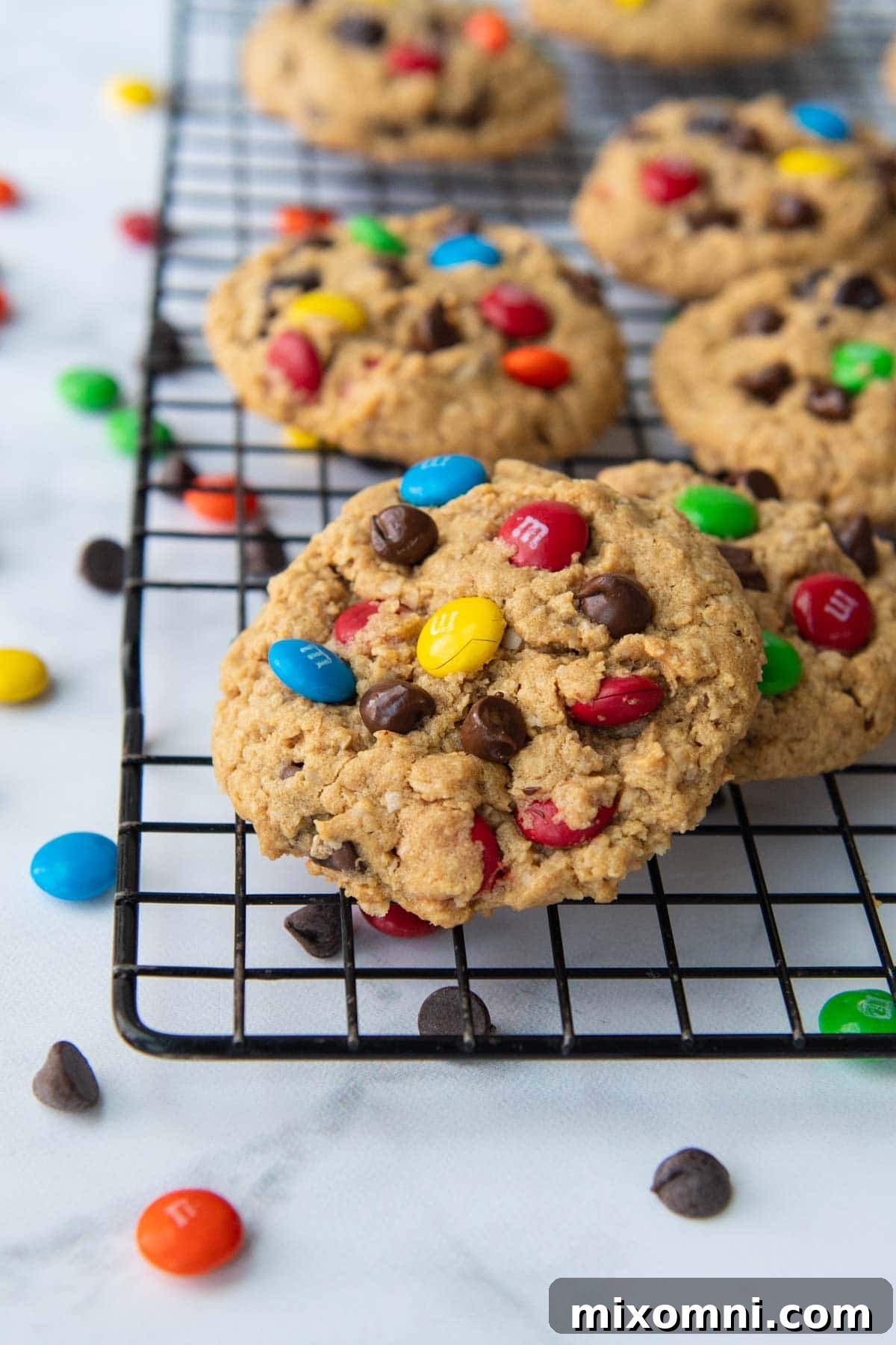A single baked gluten-free monster cookie, perfectly golden with visible M&M's and chocolate chips, resting on a black cooling rack, with another cookie blurred in the background.