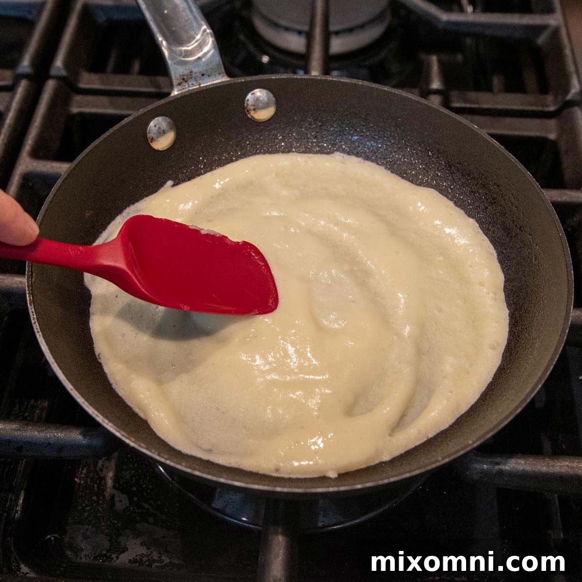 Gluten-free crepe batter being spread evenly in a hot skillet with a rubber spatula.