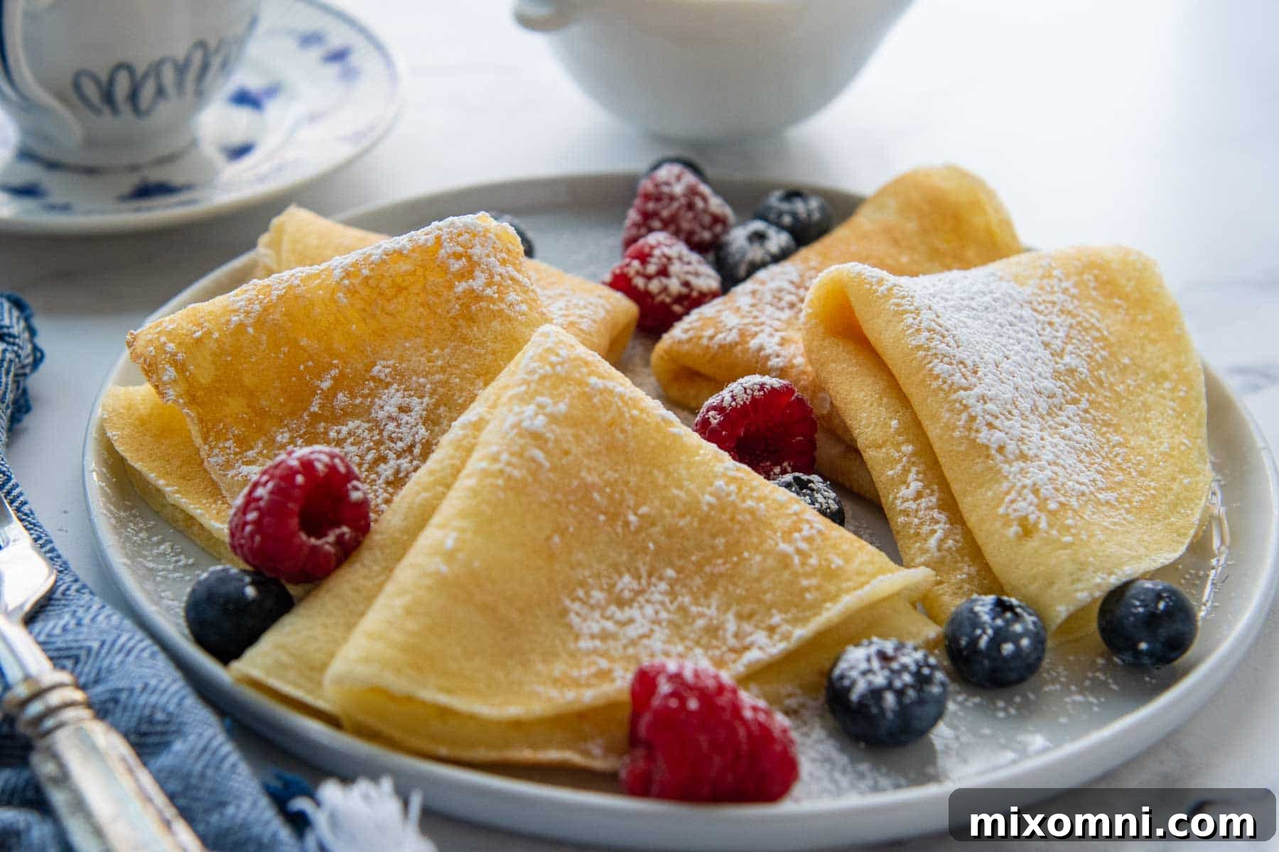 A beautifully arranged plate of golden-brown gluten-free crepes, dusted with powdered sugar and garnished with fresh raspberries and blueberries.