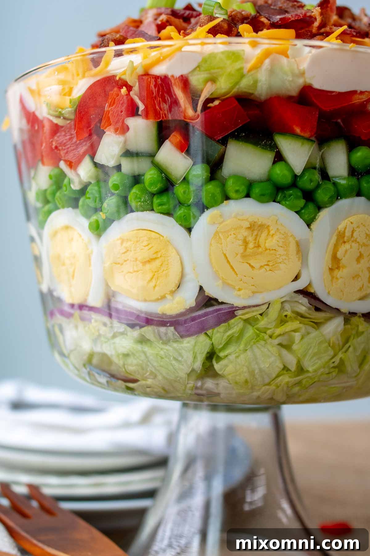 A beautiful close-up of the distinct layers in an overnight 7-layer salad, elegantly served in a clear glass trifle bowl, revealing crisp lettuce, green peas, cheese, and savory bacon.