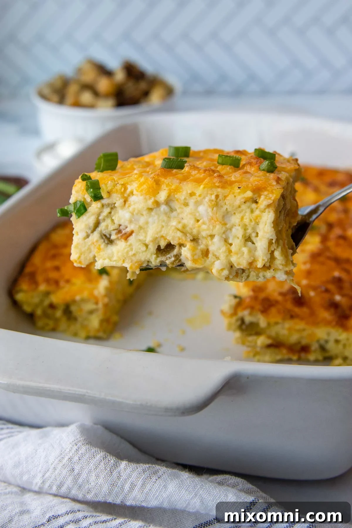 A spatula lifting out a golden-brown slice of fluffy Egg Green Chile Casserole, revealing its cheesy, eggy layers and flecks of green chili.