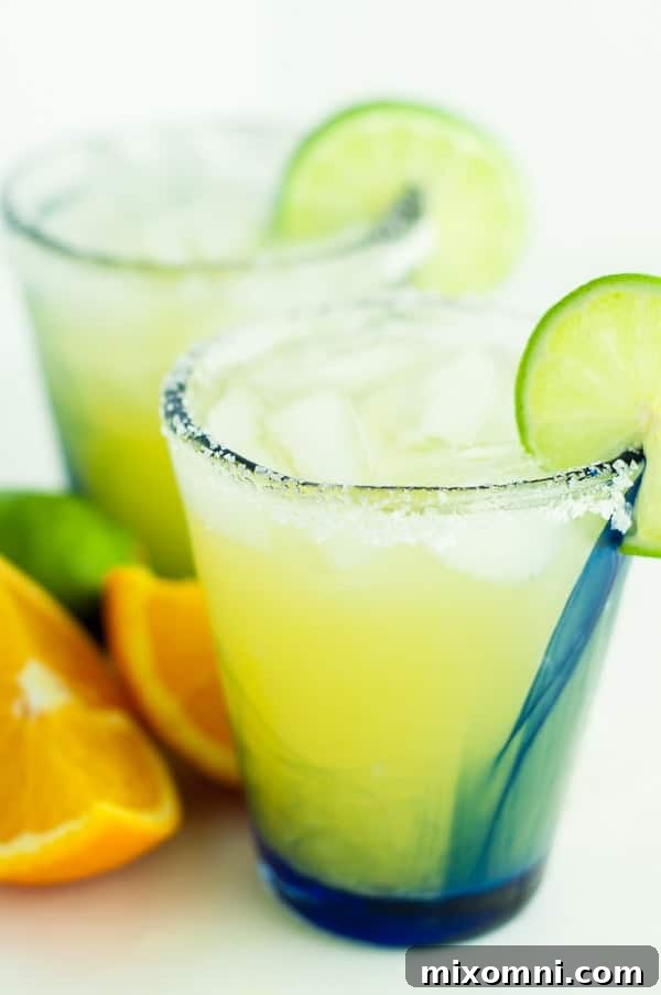 Two glasses of skinny margarita with sliced oranges and limes resting beside them on a light surface