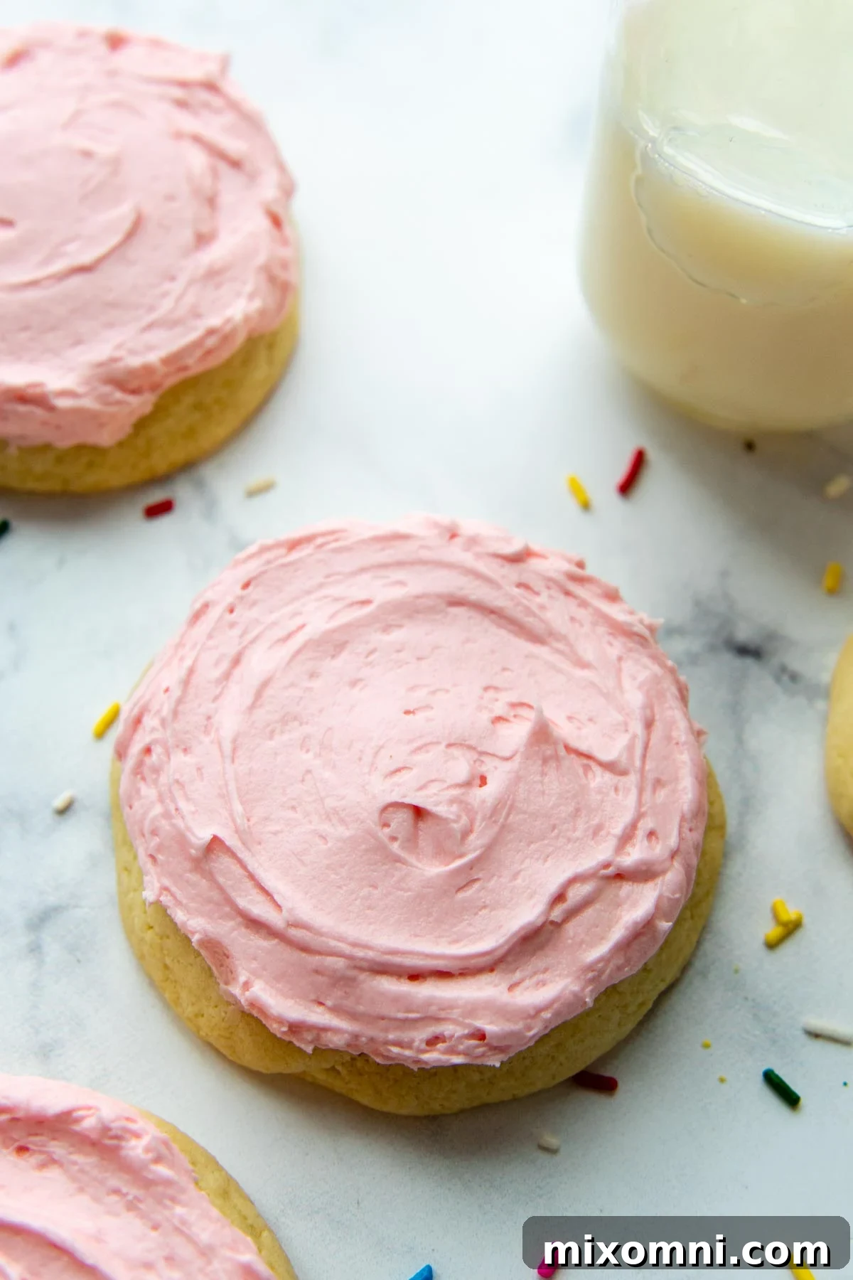 A vibrant pink sugar cookie rests on a white background, surrounded by colorful sprinkles, with a glass of milk nearby.