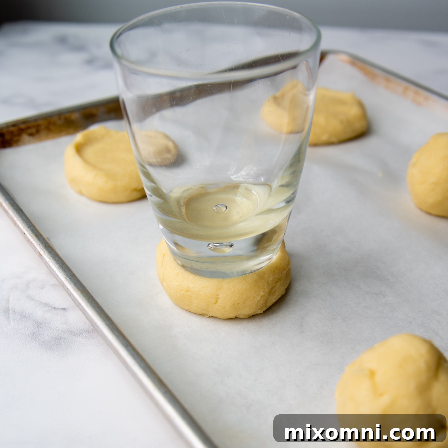A flat-bottomed glass being used to gently flatten a cookie dough ball on a baking sheet, with sugar clinging to the glass.