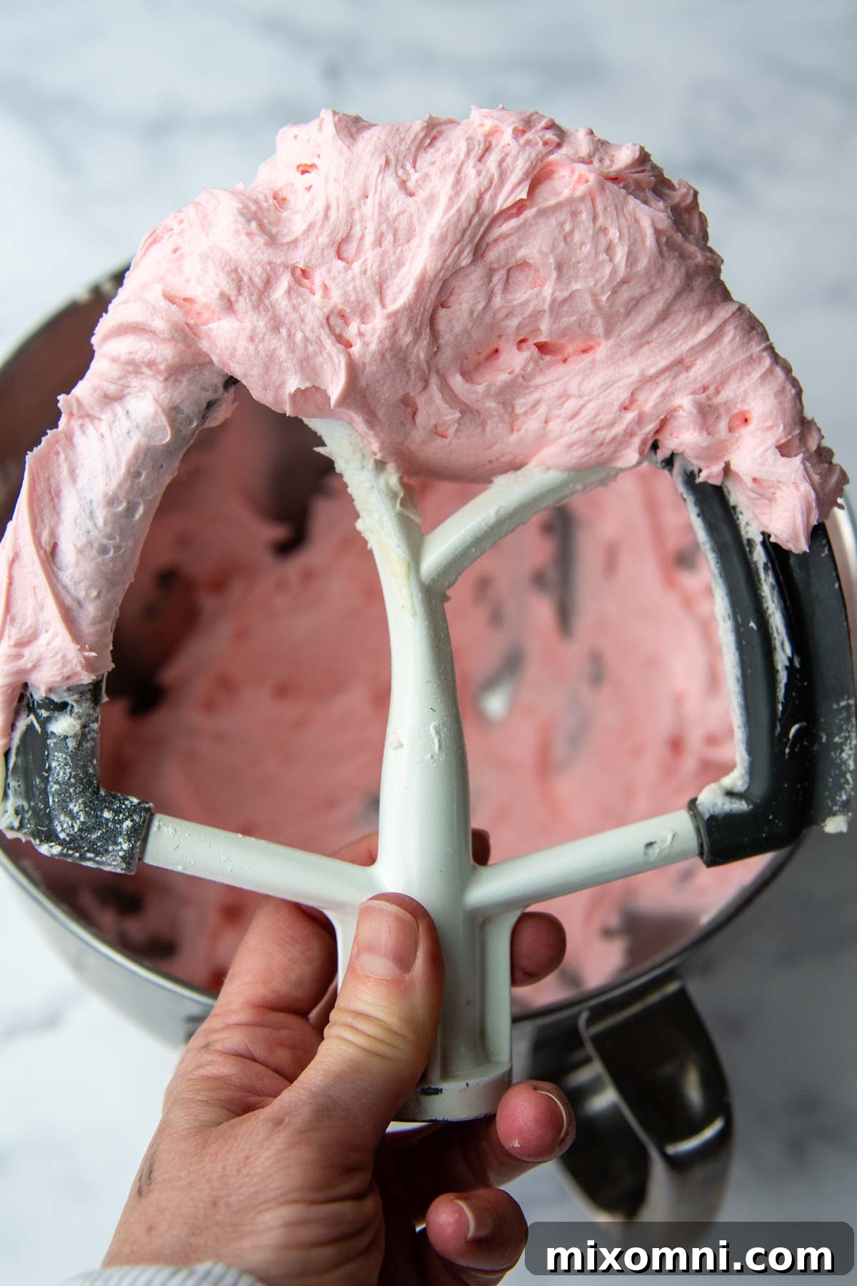 A mixer beater holding up a generous dollop of perfectly whipped, fluffy pink buttercream frosting.