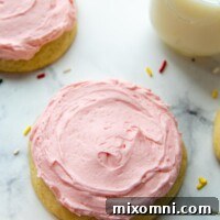 A perfectly pink sugar cookie on a white background, surrounded by sprinkles, with a glass of milk.
