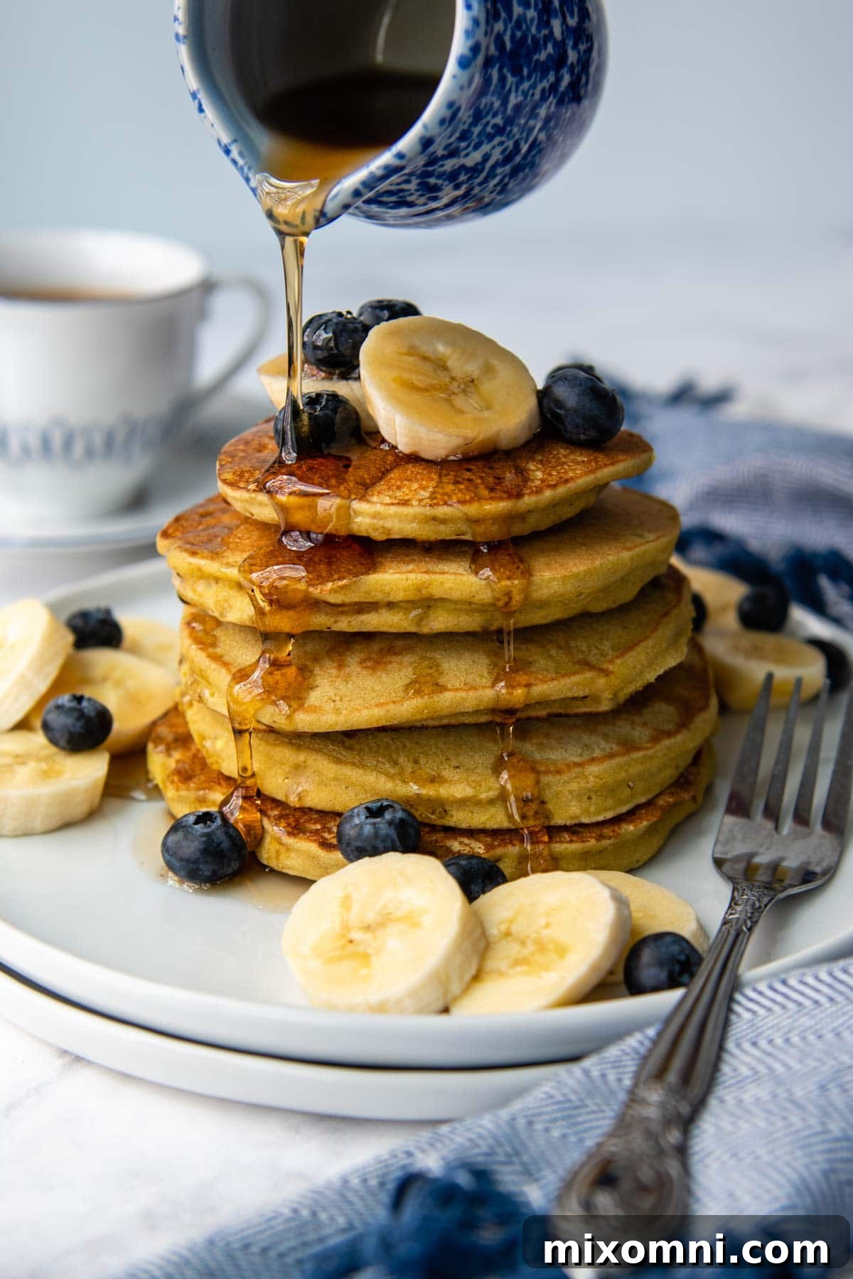 Syrup being poured on a stack of golden brown banana buckwheat pancakes, garnished with fresh slices of banana and vibrant blueberries.