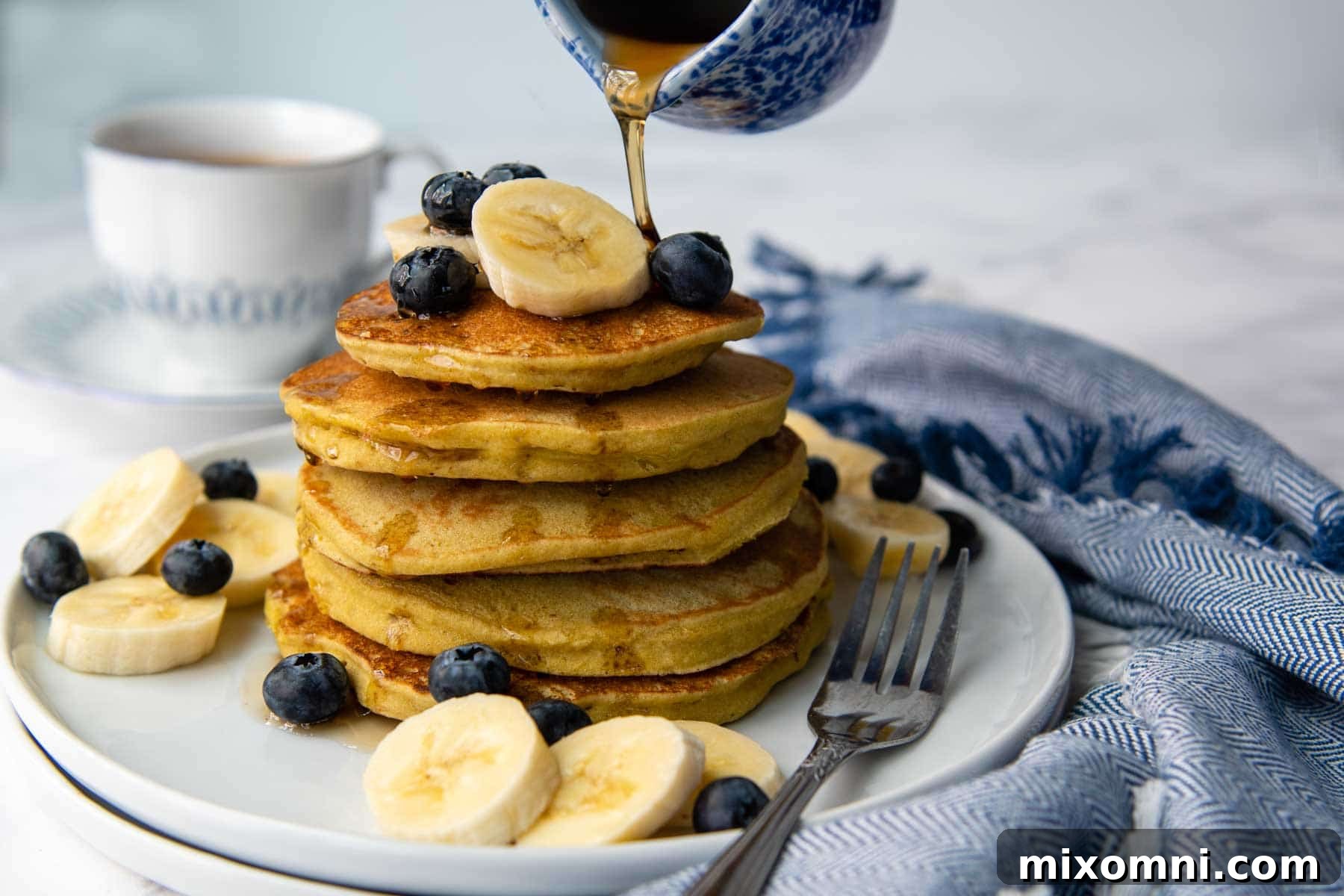 A small, elegant blue carafe pouring golden syrup generously over a delightful stack of fluffy buckwheat banana pancakes.