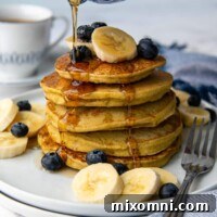 Syrup being poured on a stack of pancakes with bananas and blueberries scattered around.