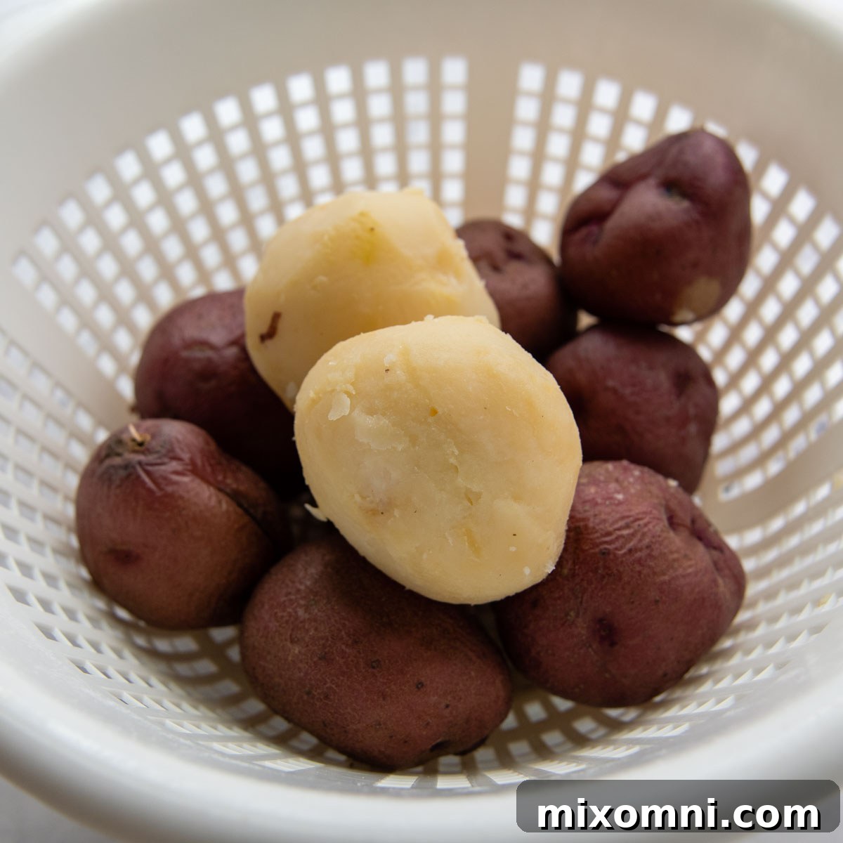 A colander filled with perfectly boiled red potatoes, with one potato peeled to show its tender interior, ready for a delicious potato salad.