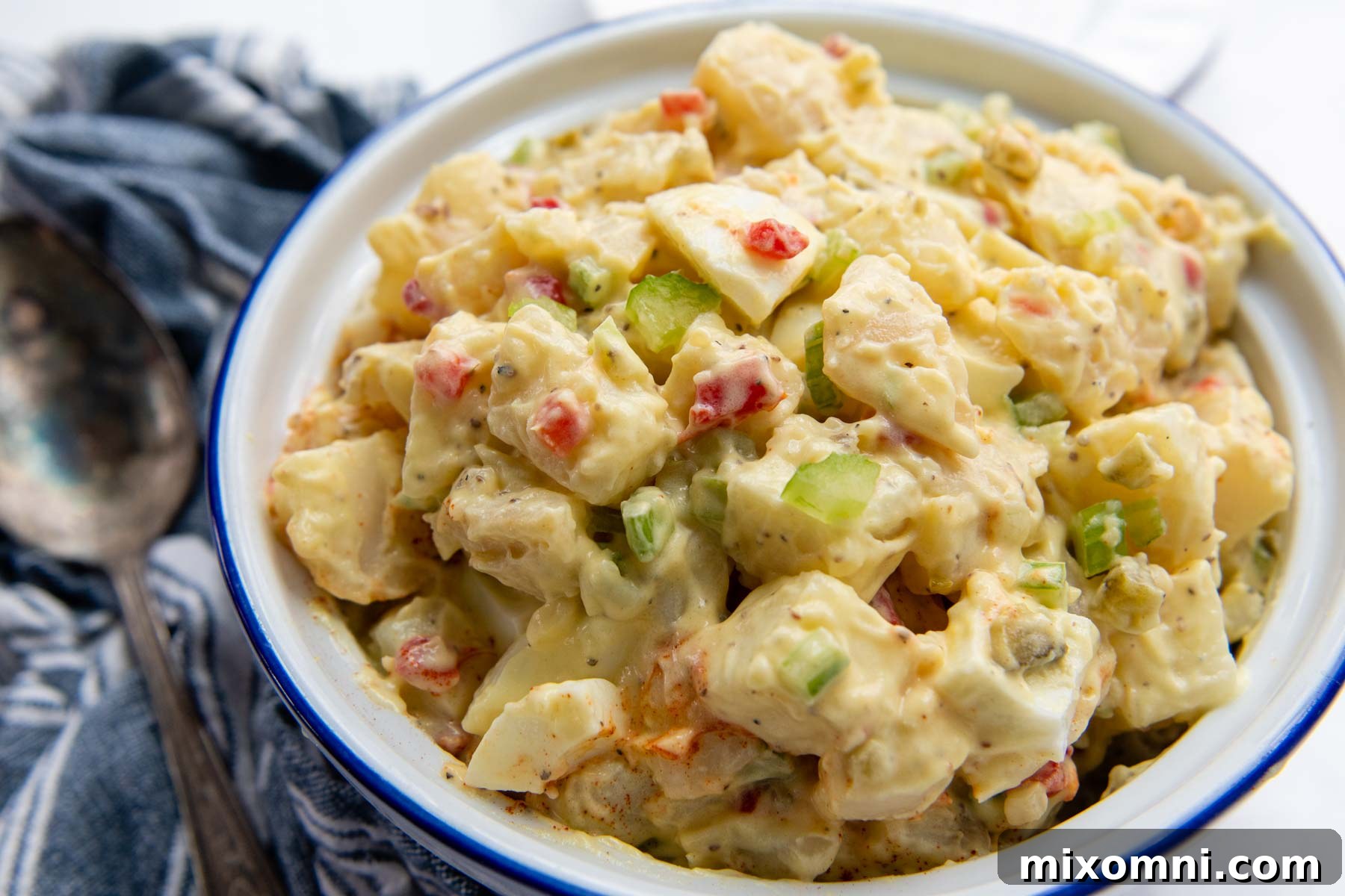 A horizontal view of a vibrant bowl of Amish potato salad, garnished with a sprinkle of paprika and fresh chives, set on a rustic blue linen next to a wooden serving spoon.