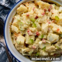 An inviting overhead shot of delicious homemade Amish potato salad in a white ceramic bowl, garnished with fresh parsley, sitting on a wooden surface with a rustic blue linen napkin beside it.