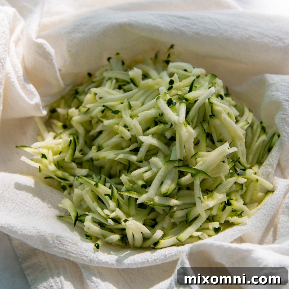 Grated zucchini being squeezed in a white kitchen towel to remove excess moisture.