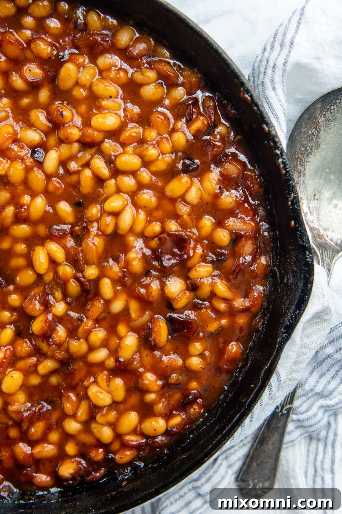 Overhead shot of baked beans in a cast iron skillet with a spoon next to it, showcasing their rich texture.