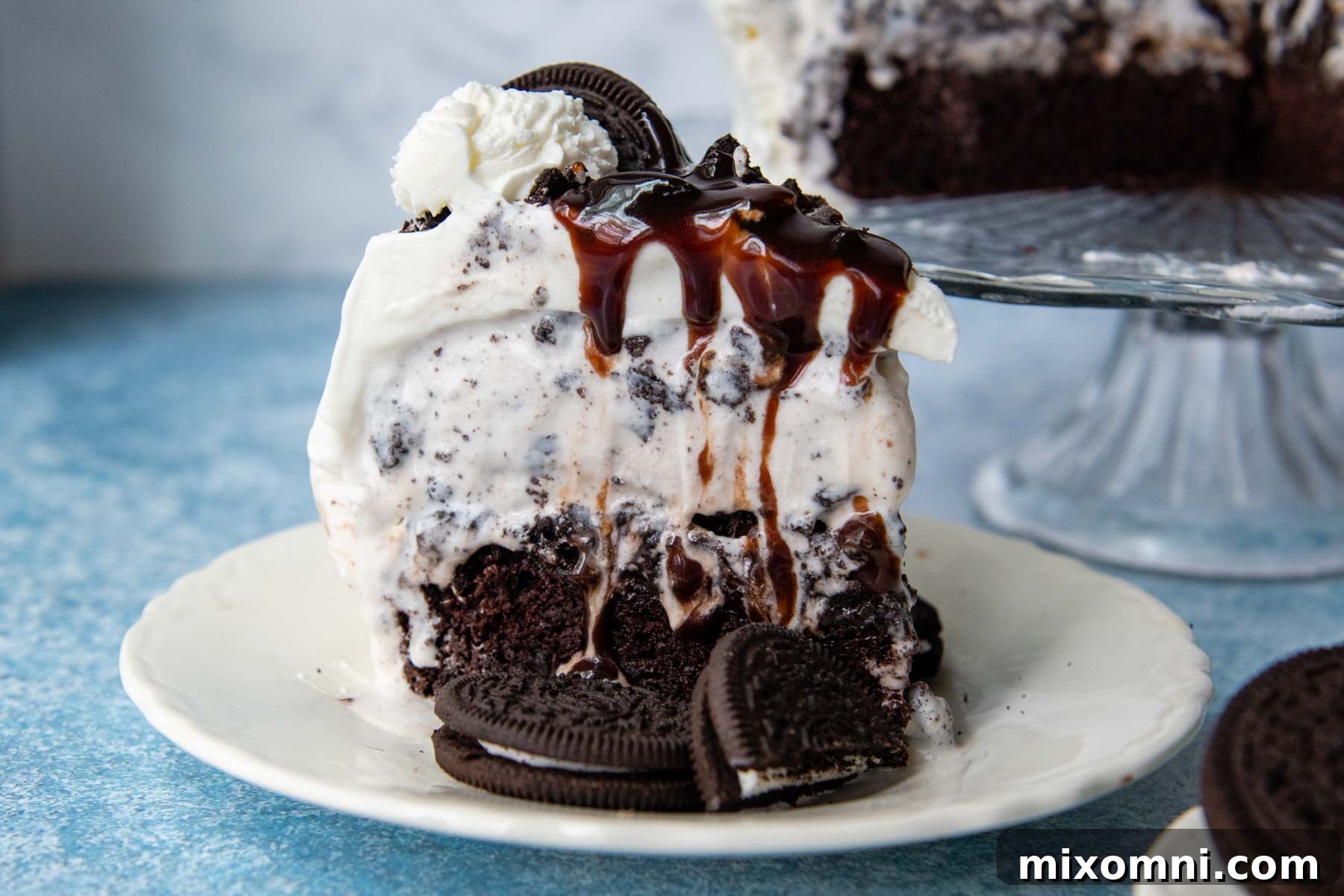 A beautifully presented slice of ice cream cake on a white plate, garnished with crushed Oreos and a drizzle of hot fudge, ready to be enjoyed.