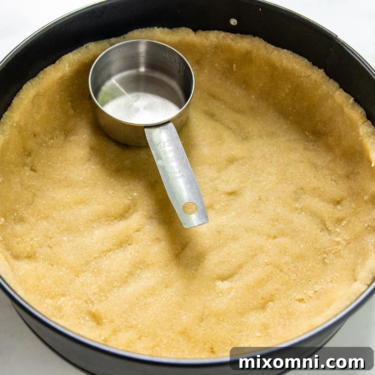 A measuring cup pressing the almond flour crust firmly into a springform pan.