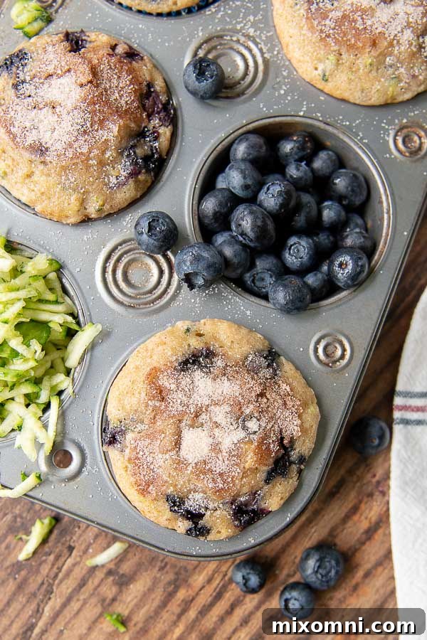 Close-up shot of individual blueberry zucchini muffins baked in a tin, with extra blueberries and shredded zucchini in the background, hinting at their delicious ingredients.