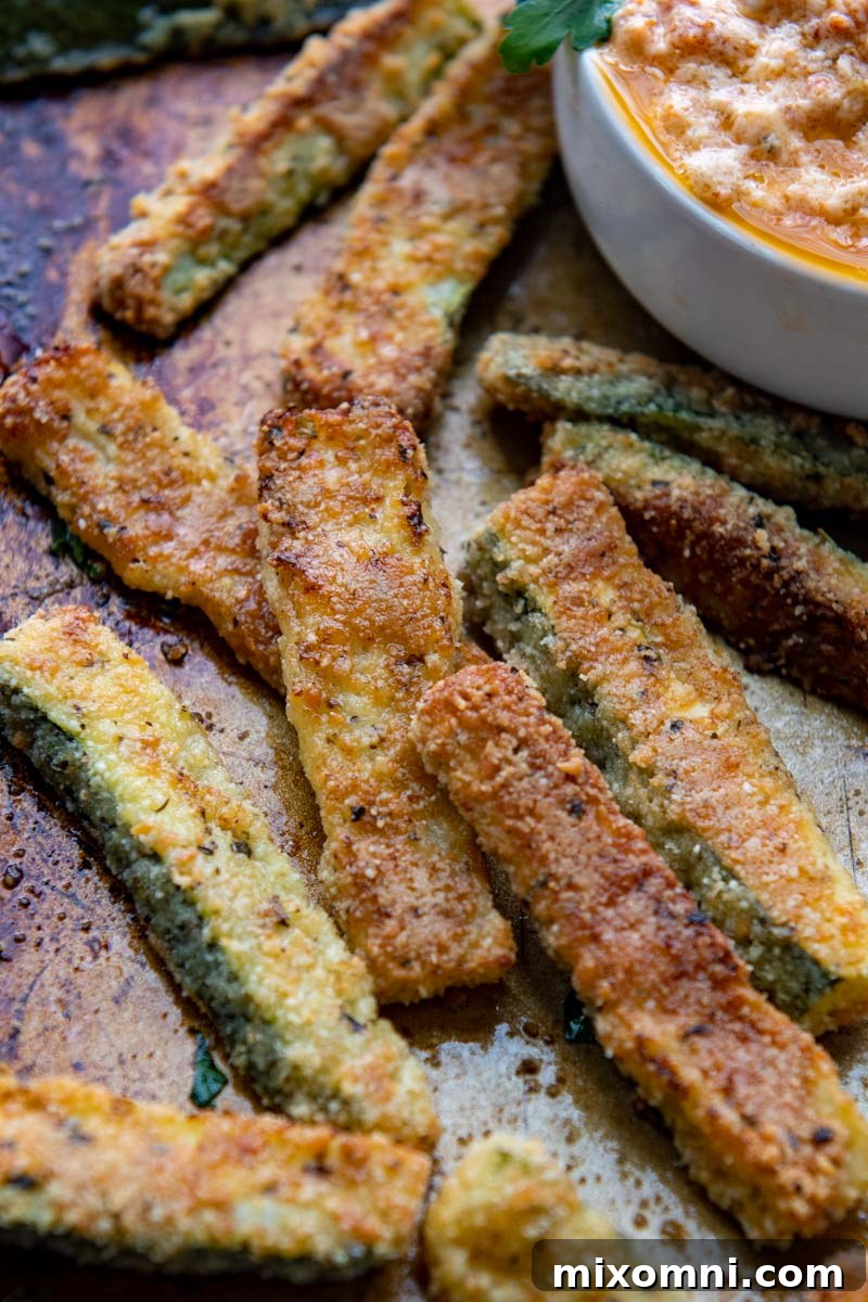 Close up of baked zucchini fries on a baking sheet with a dipping sauce in the background.