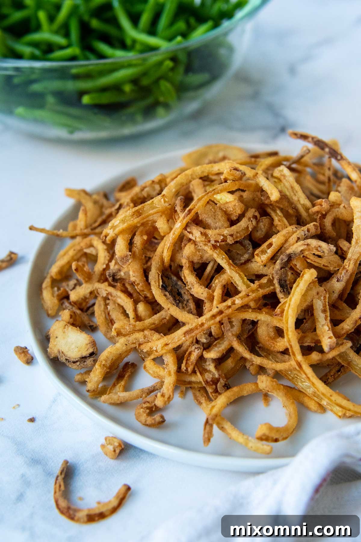 A plate piled high with crispy gluten-free French fried onions, with a bowl of green beans blurred in the background.