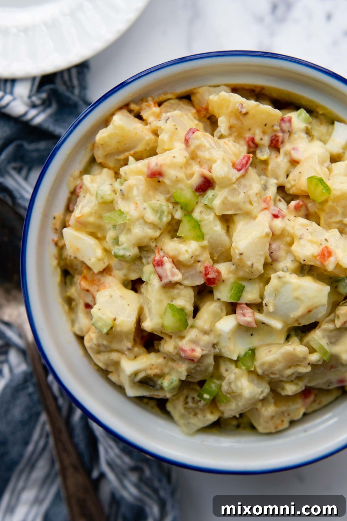 Overhead shot of creamy Amish potato salad in a white serving bowl, with a charming blue linen napkin next to it.