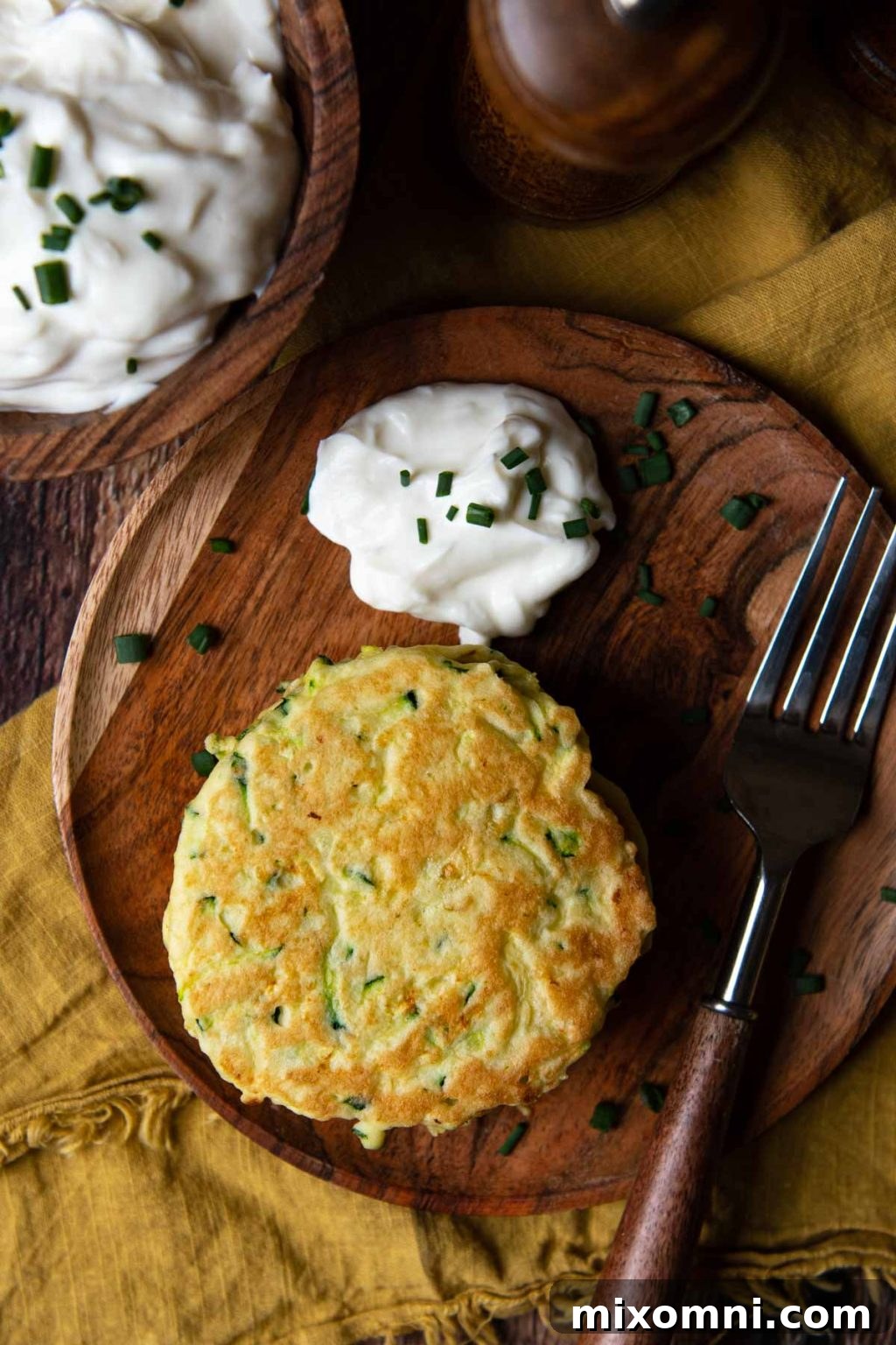 Overhead shot of golden-brown gluten-free zucchini pancakes on a rustic wood plate, garnished with a dollop of sour cream and fresh herbs.