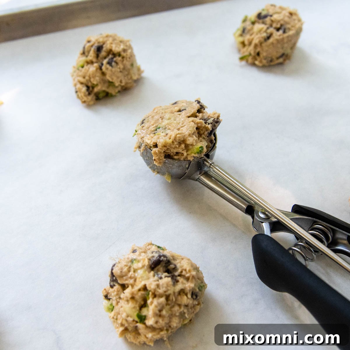 A cookie scoop placing rounded portions of raw gluten-free zucchini cookie dough onto a parchment-lined baking sheet, ready for the oven.