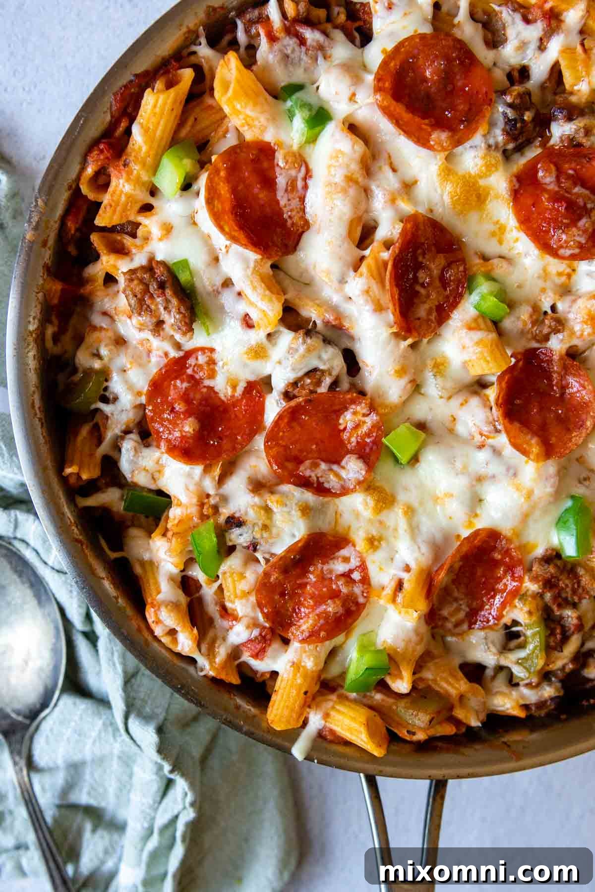 Close-up of a bubbling, cheesy Supreme Pizza Pasta Skillet, ready to serve, with a spoon and linen next to it.