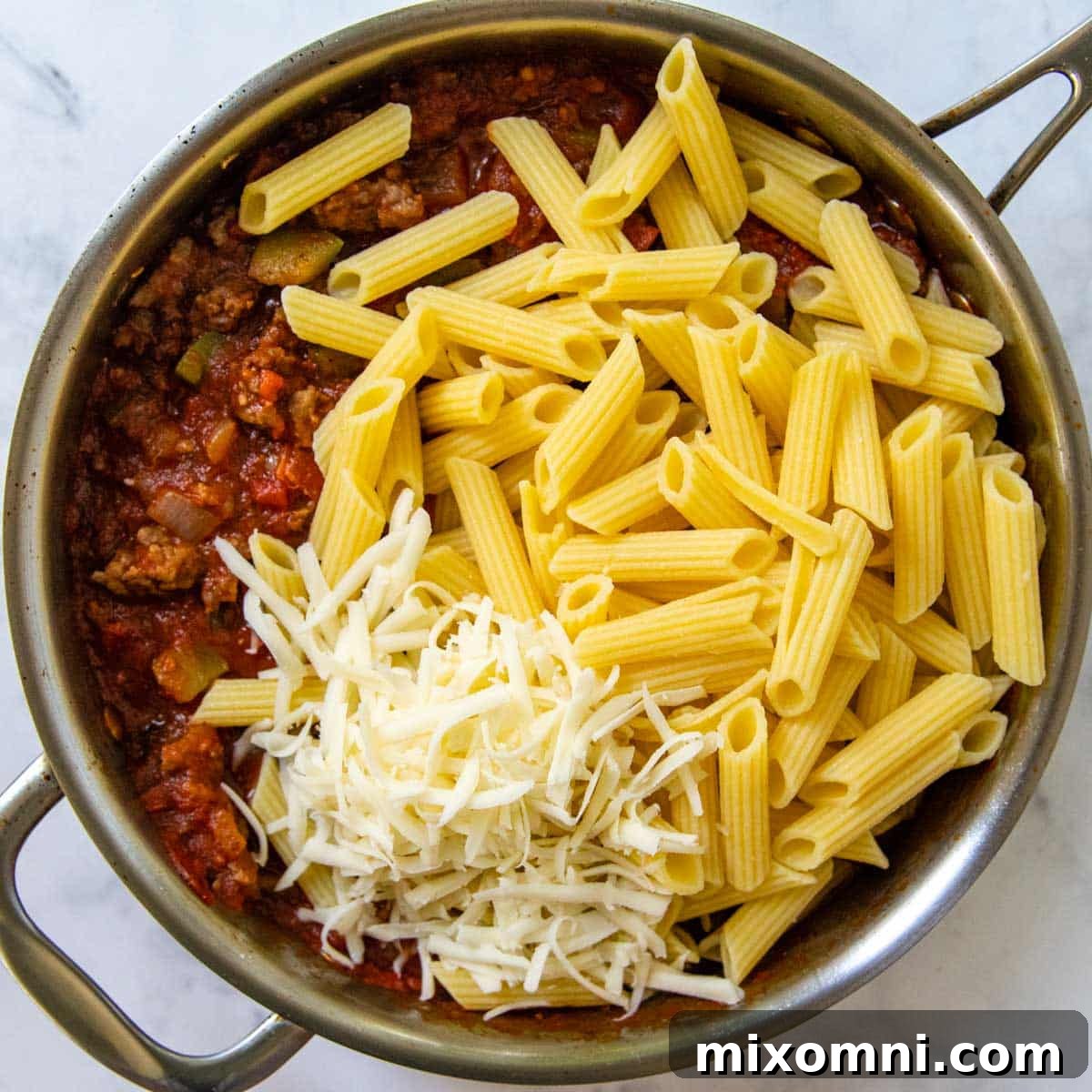 The perfectly cooked pasta being mixed into the rich, simmering pizza sauce with sausage and vegetables in a skillet.