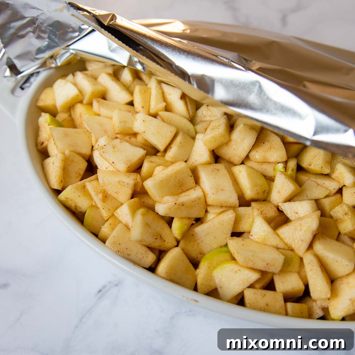 Unbaked apples in a baking dish, tightly covered with aluminum foil, ready for their initial bake.