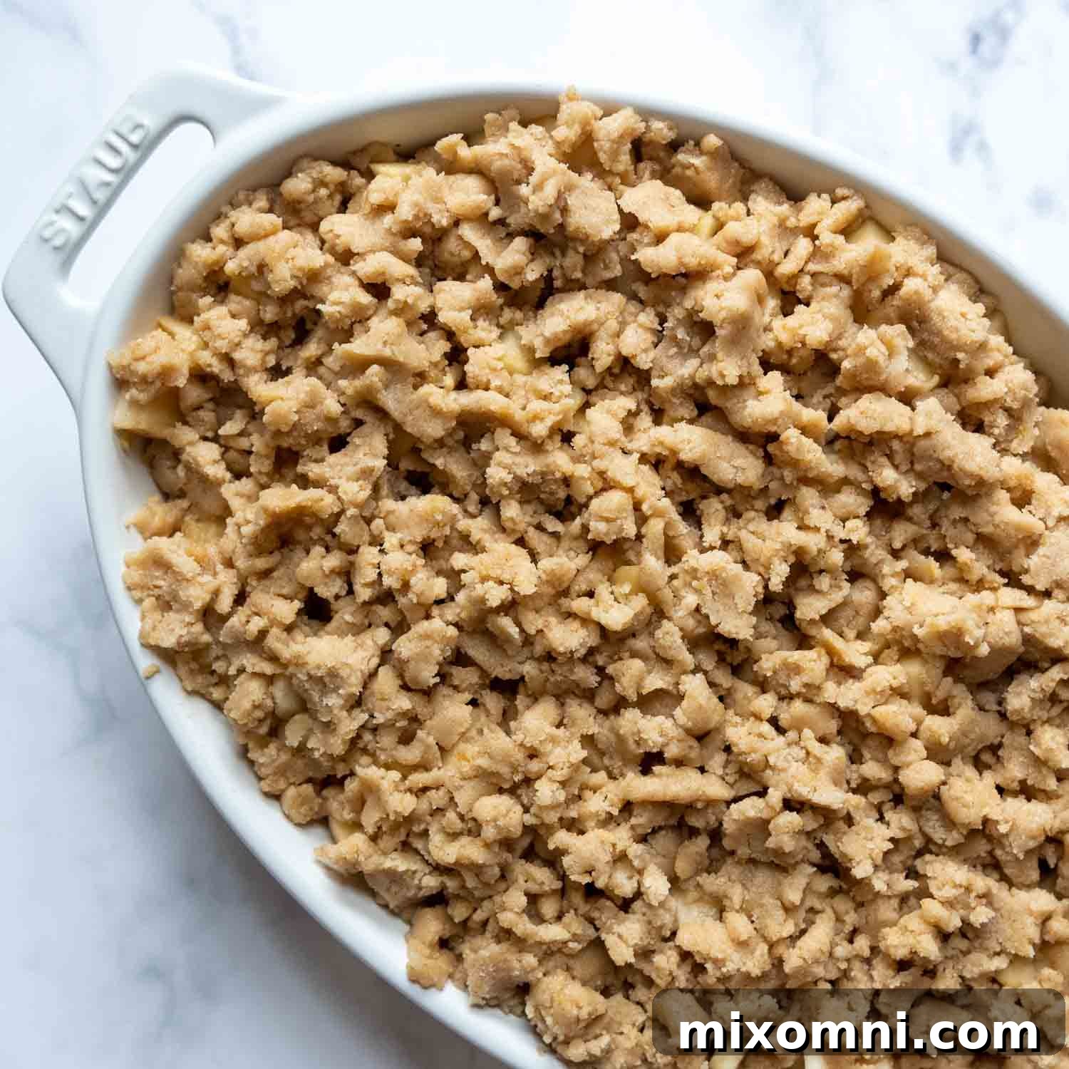 Unbaked apple crisp in a white oval baking dish, topped with generous streusel crumbles, ready for the final bake.