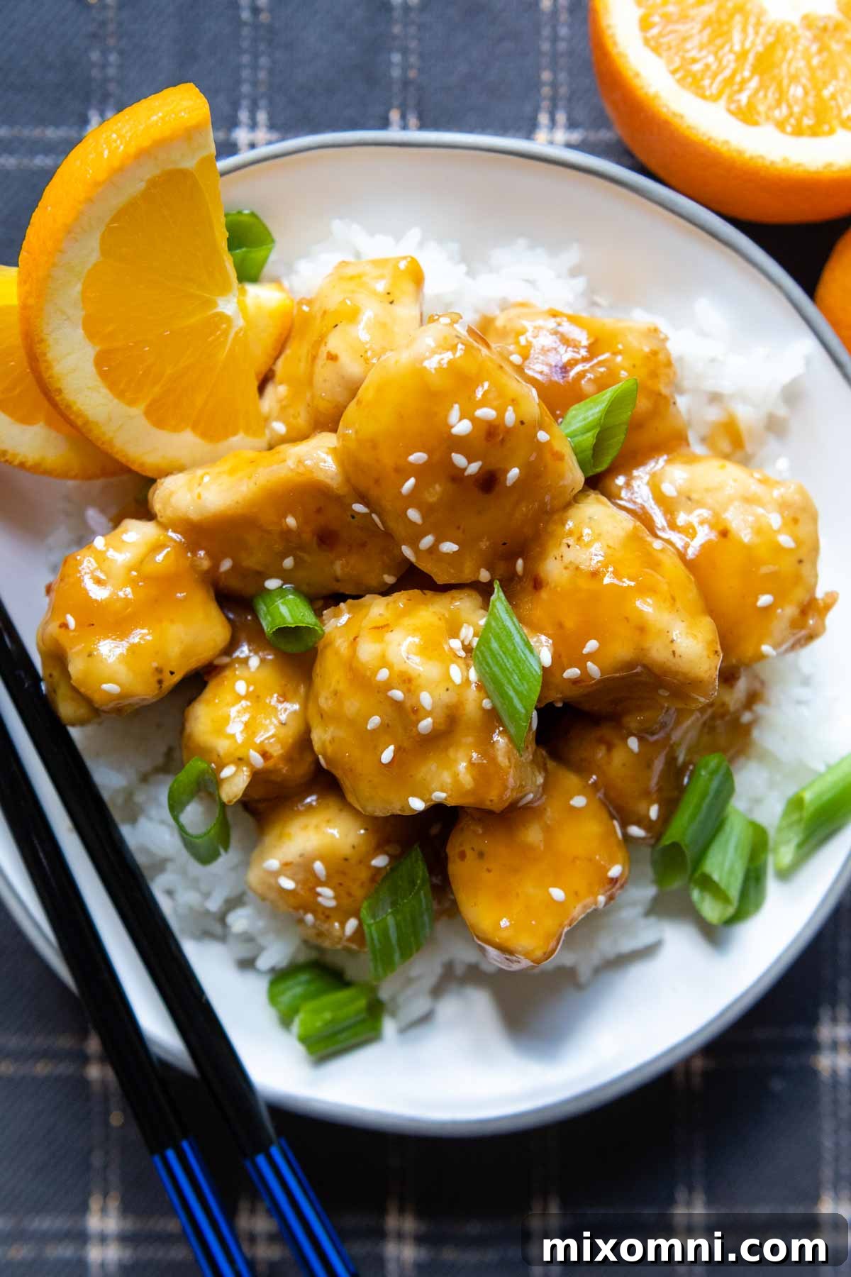 Overhead shot of a serving of crispy gluten-free orange chicken in a white bowl with chopsticks, garnished with fresh orange slices and green onions.