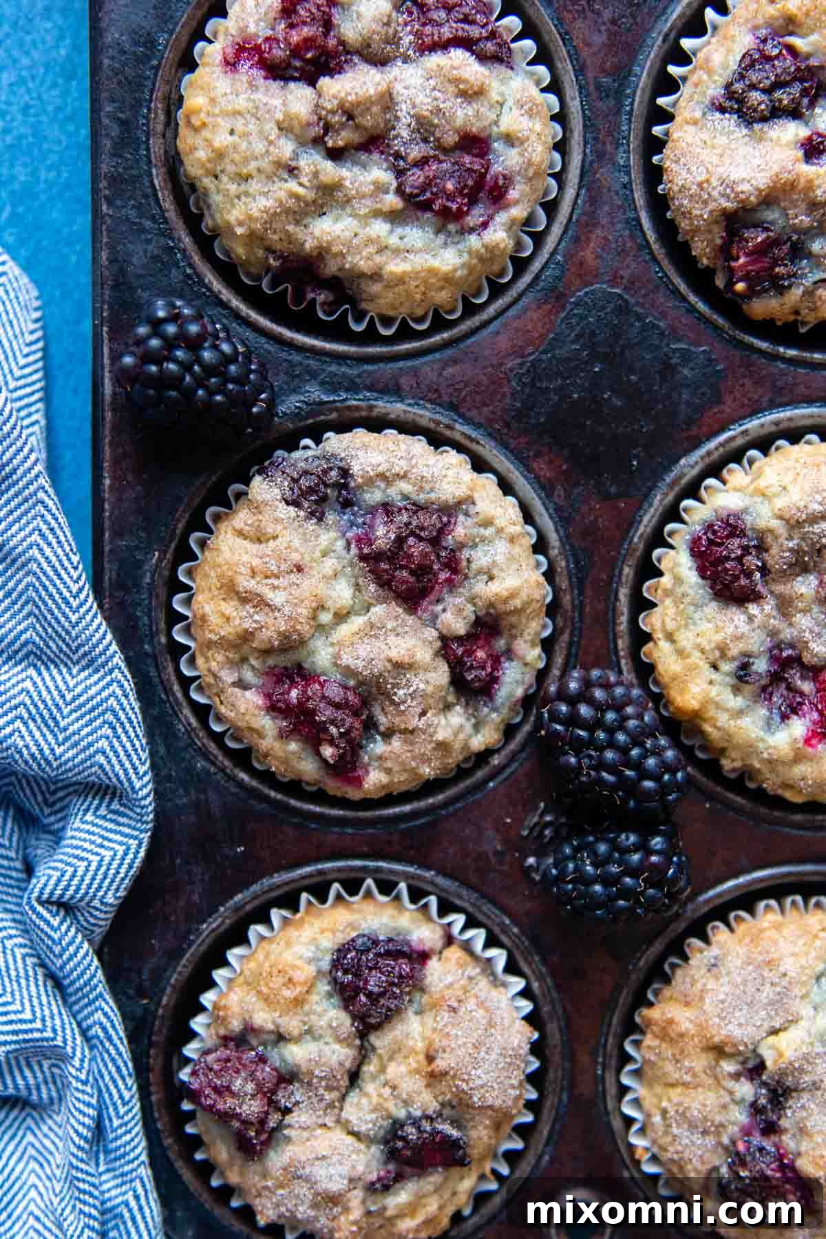 Overhead shot of freshly baked gluten-free blackberry muffins nestled in a dark muffin tin, with a soft blue kitchen towel beside it, evoking a cozy home baking scene.