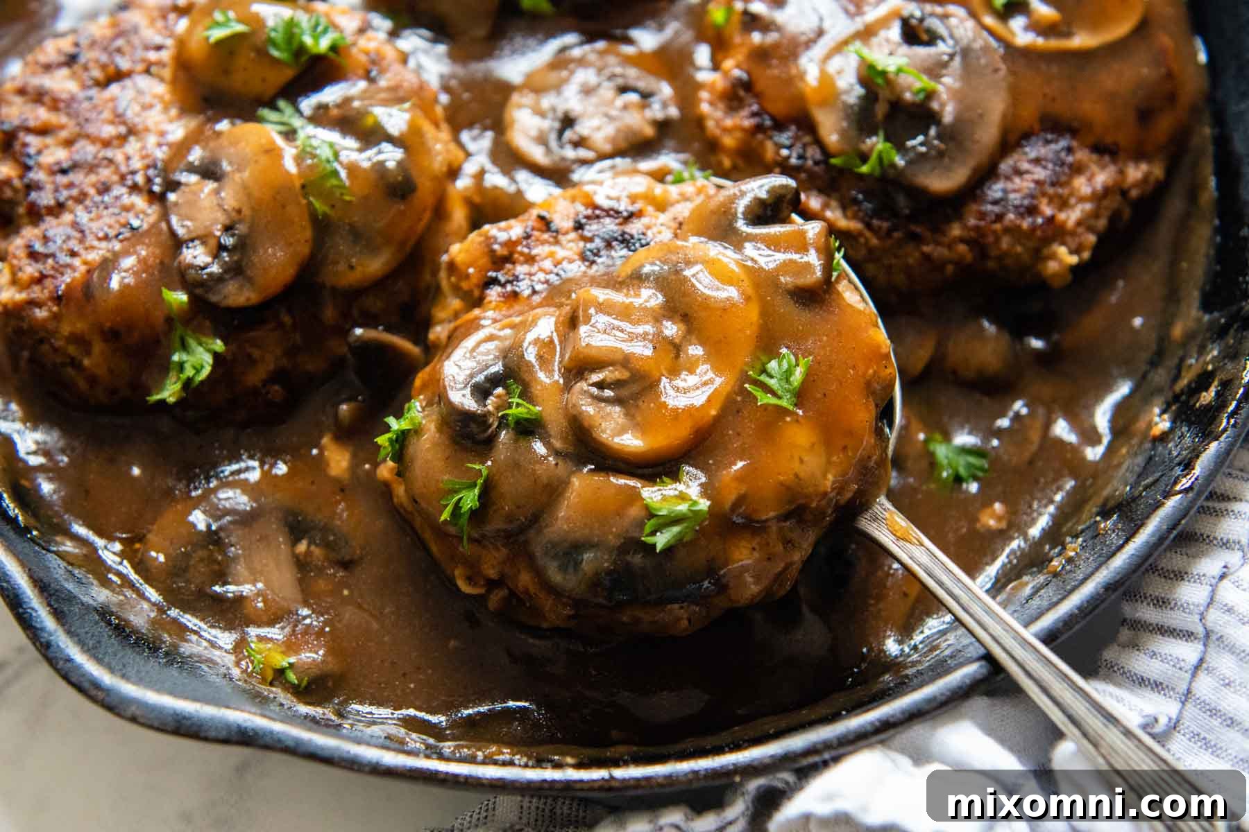 A close-up of a spoon lifting a Salisbury steak topped with mushrooms and rich gravy from a skillet.