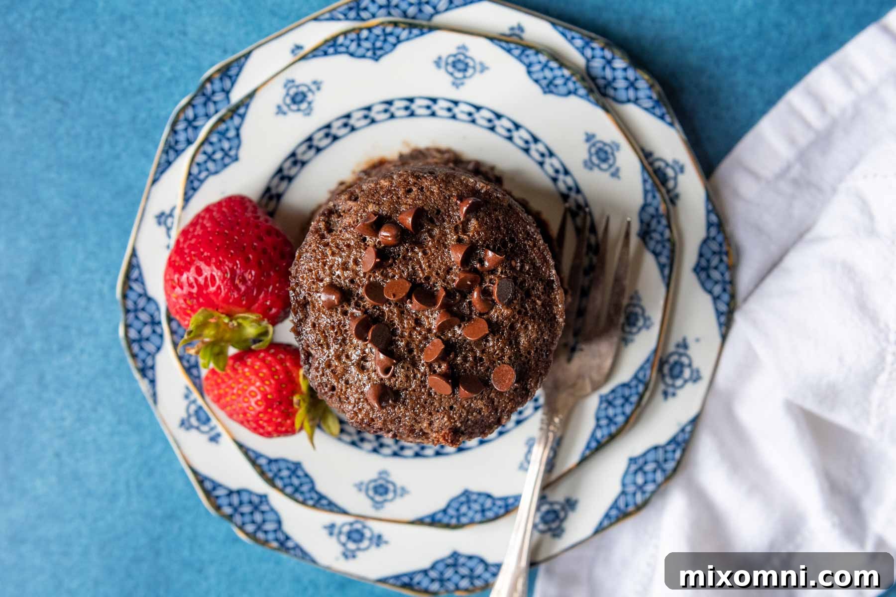 An overhead shot showcasing the top of a beautifully baked chocolate almond flour mug cake, artfully presented on a white and blue plate, accompanied by fresh strawberries.