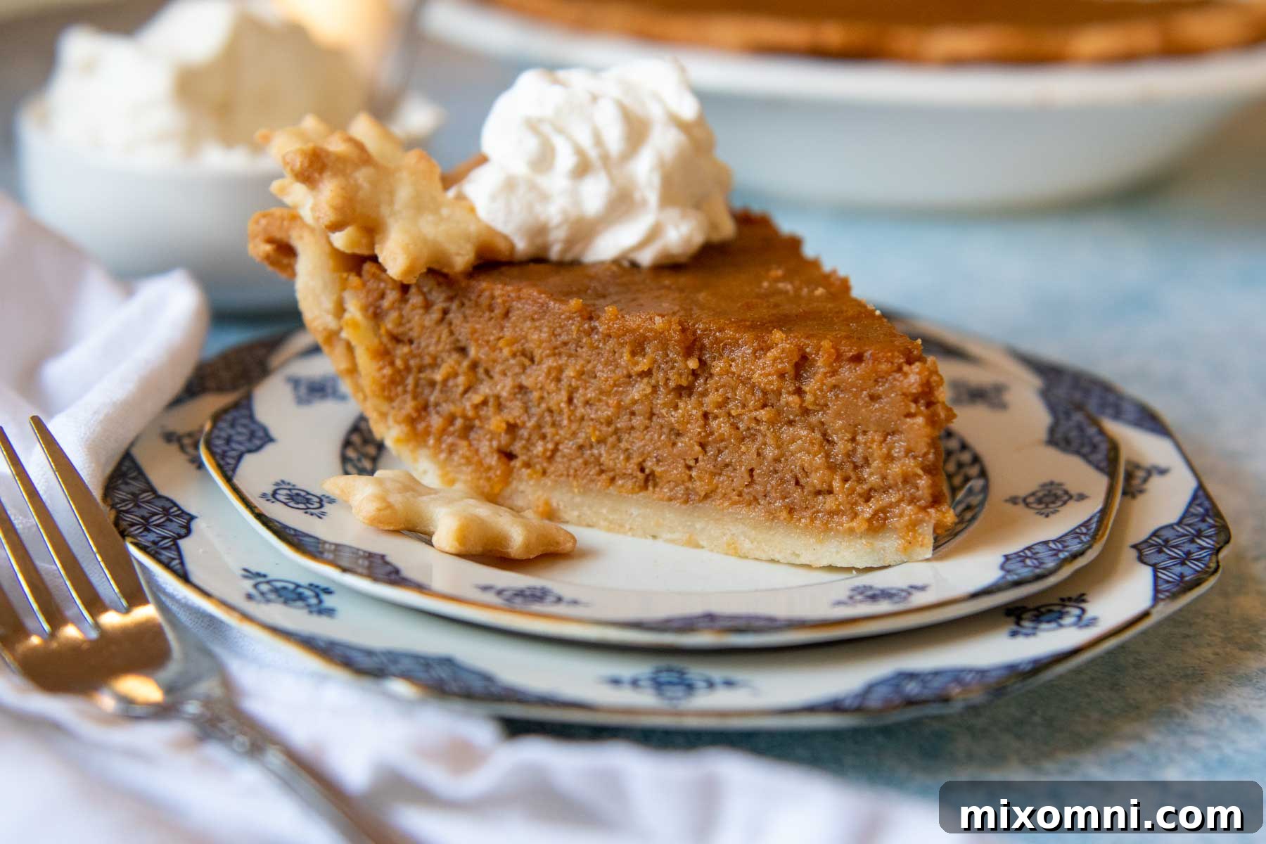 A slice of gluten-free sweet potato pie topped with a generous swirl of whipped cream on a white plate, with the whole pie blurred in the background, ready for serving.