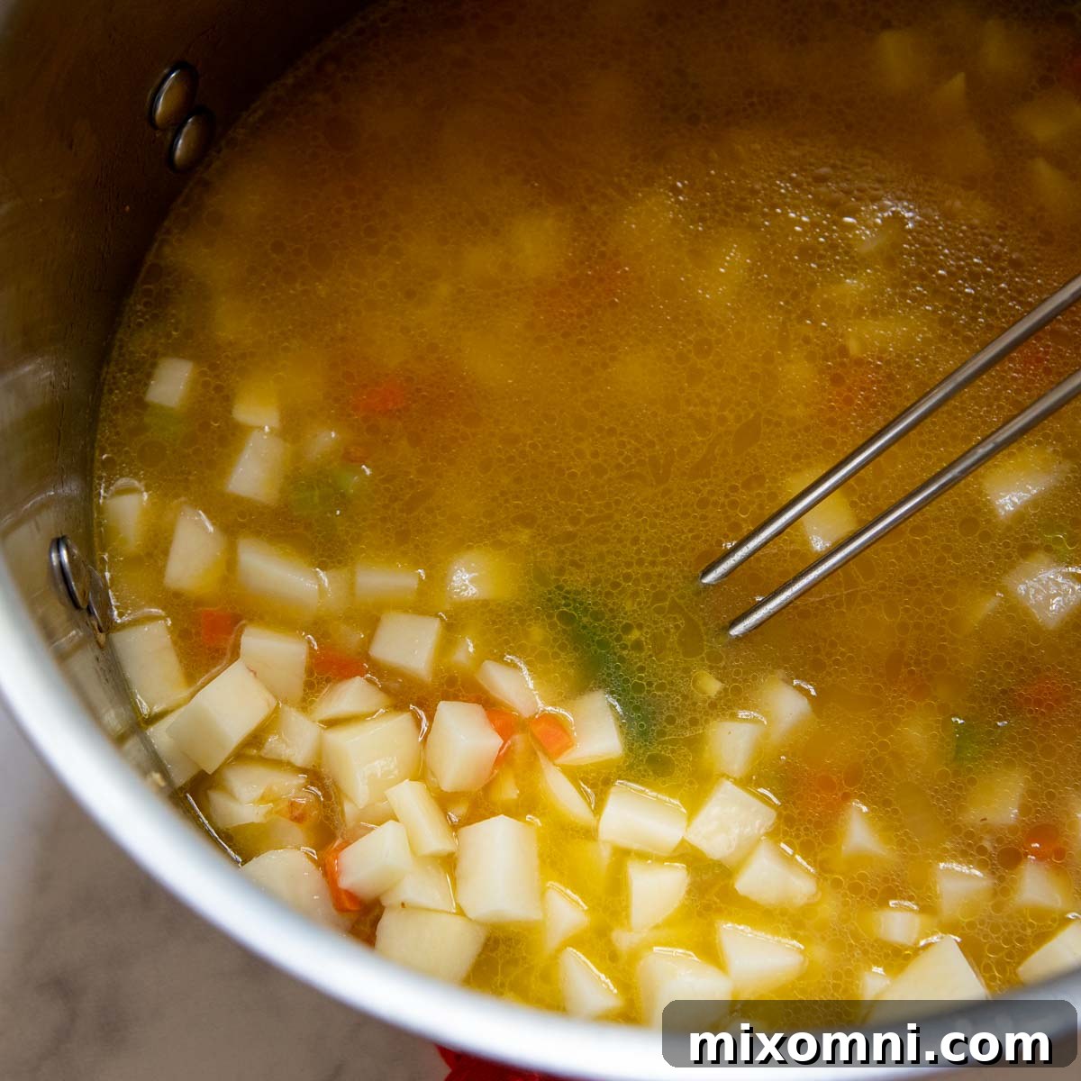 A large pot of potato soup simmering gently on the stovetop, with tender potato cubes and vegetables visible, being stirred to ensure even cooking.