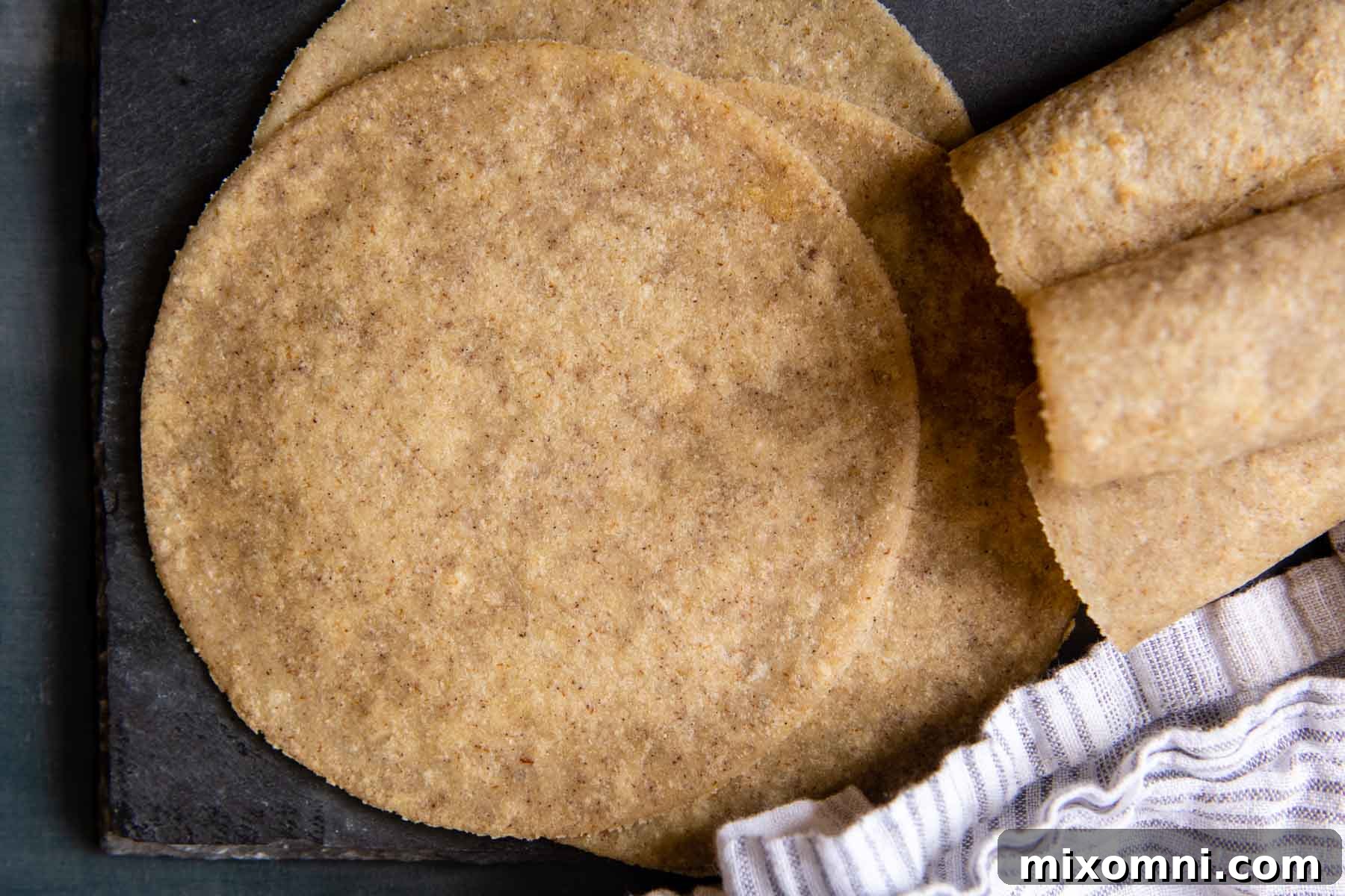 Unrolled tortillas on a dark surface next to a stack of rolled ones, ready for meal prep.